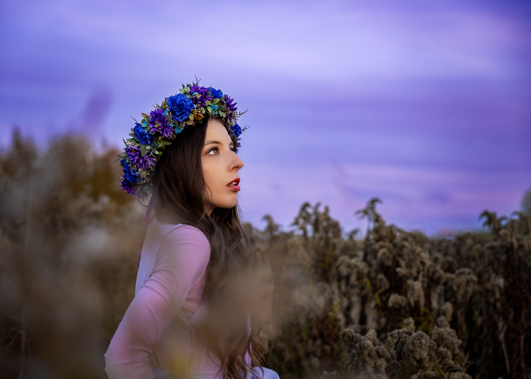 Kobieta z wiankiem z niebieskich i fioletowych kwiatów, spoglądająca w górę na tle rozmytego, fioletowego nieba i jesiennych traw.