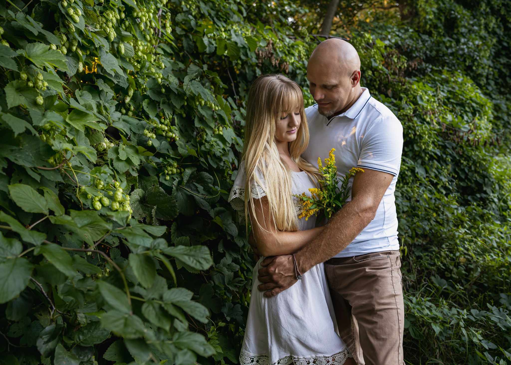 Przyszła mama w białej sukience, obejmowana przez partnera trzymającego żółte kwiaty na tle ściany z zielonego bluszczu z owocami.