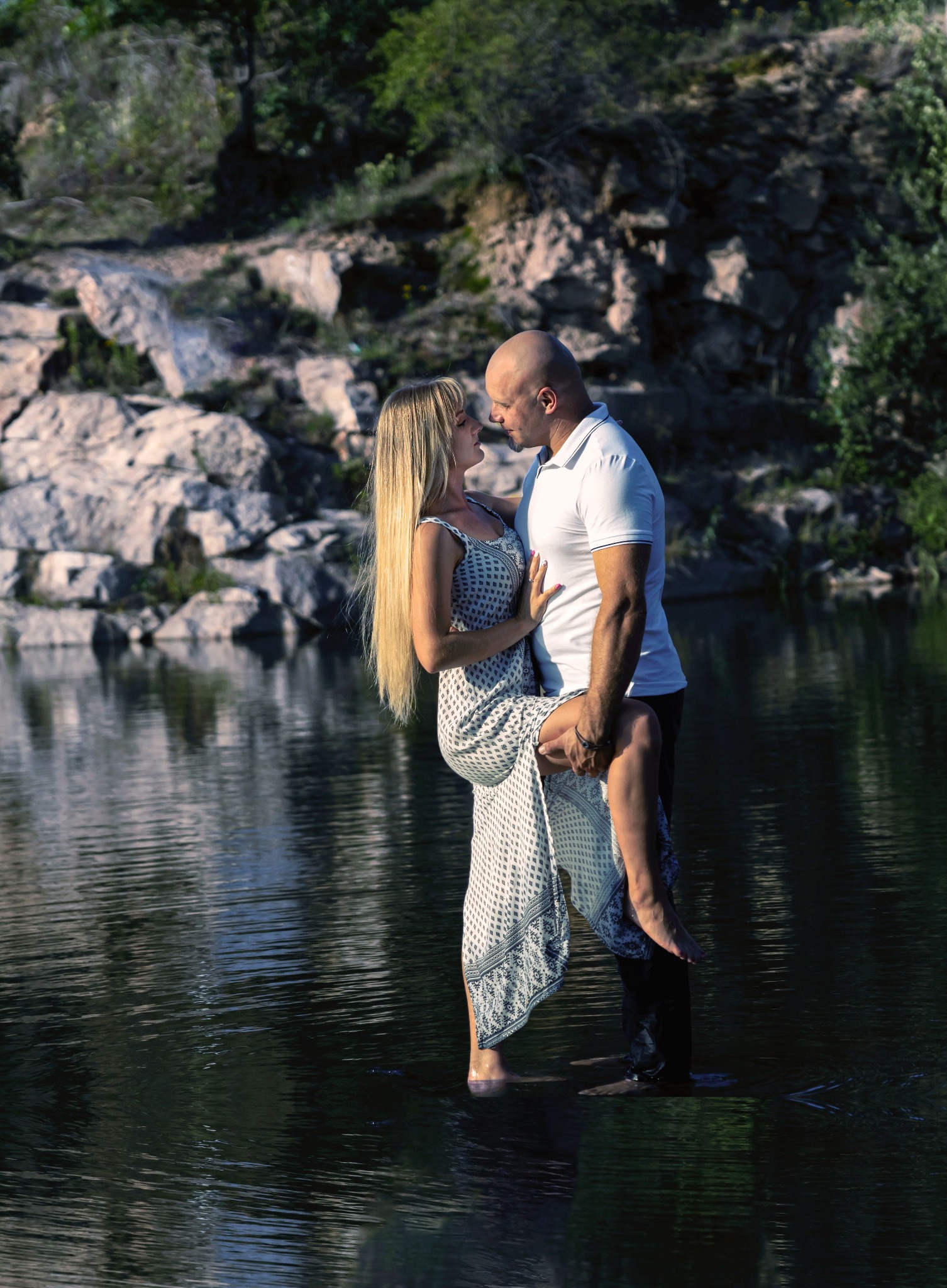 Mężczyzna trzymający kobietę w kwiecistej sukience na rękach, stojąc w wodzie, z kamienistym brzegiem w tle.