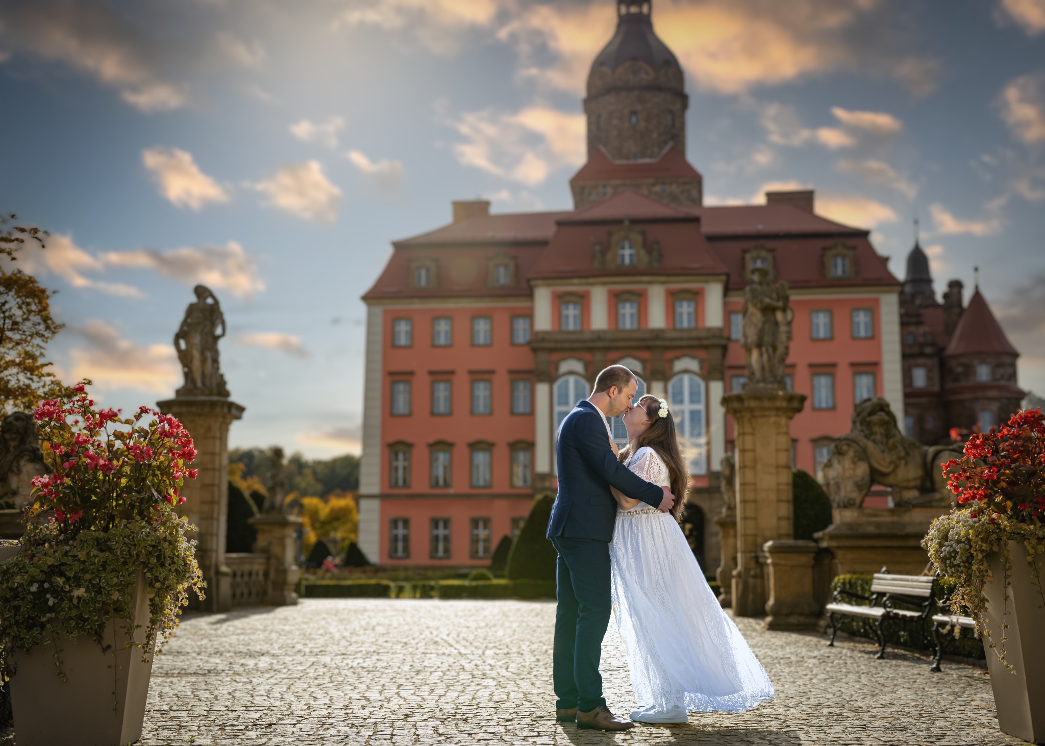 Para młoda w objęciach na tle zamku w Chróstniku, kobieta w białej sukni z koronki i wiankiem, mężczyzna w granatowym garniturze, kamienna nawierzchnia, posągi i kwiaty w donicach.