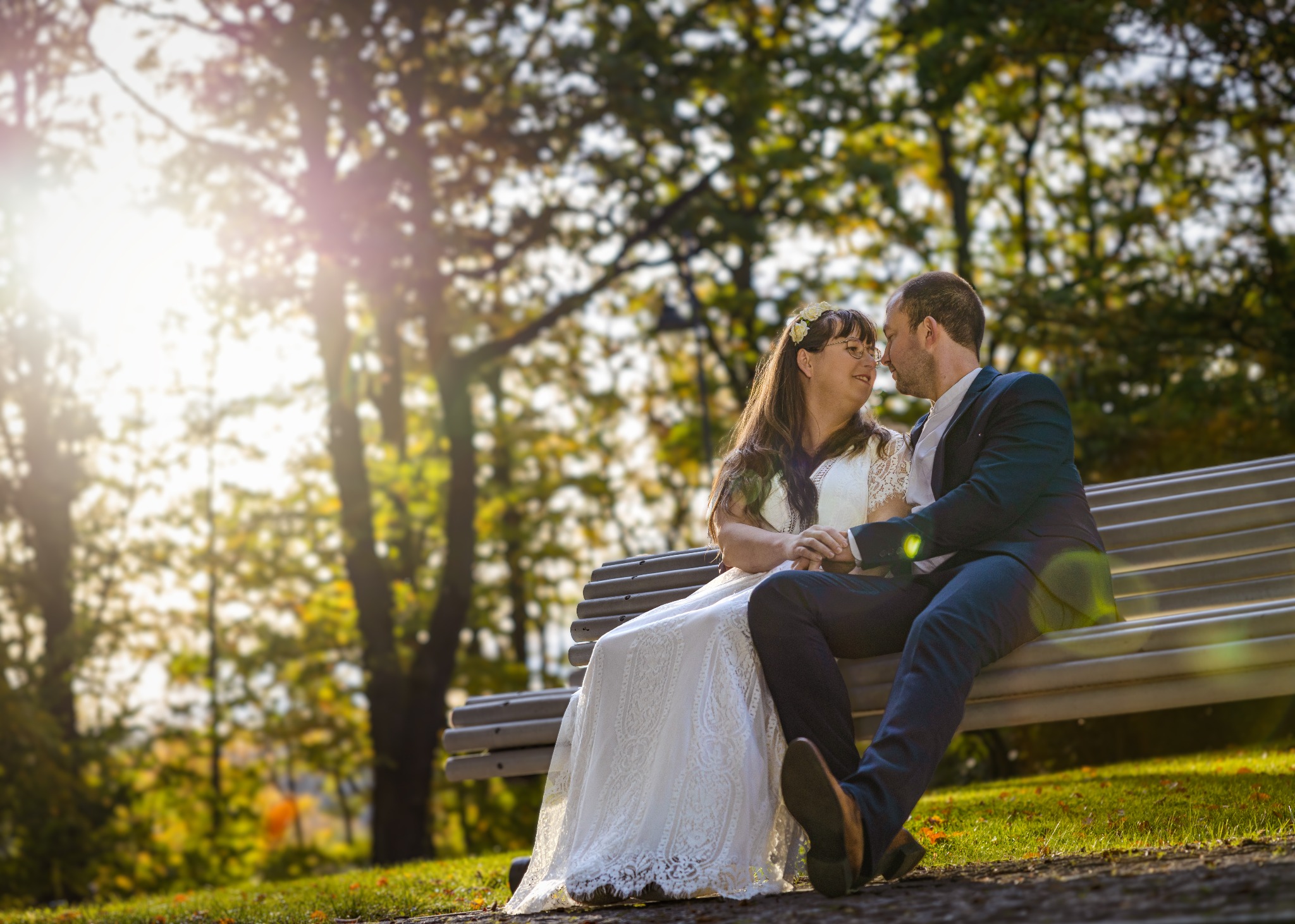 Para w strojach ślubnych siedzi na ławce w parku, mężczyzna obejmuje kobietę, w tle drzewa i promienie słońca.