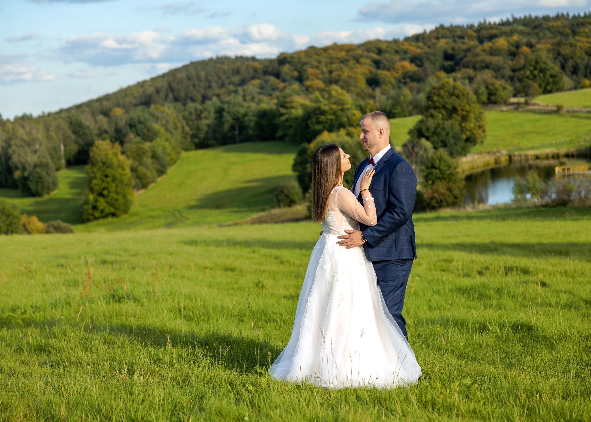Para młoda w plenerze: panna młoda w białej sukni z długimi rękawami i pan młody w granatowym garniturze w kratę, stoją na zielonej łące z widokiem na zalesione wzgórza i małe jezioro w tle...