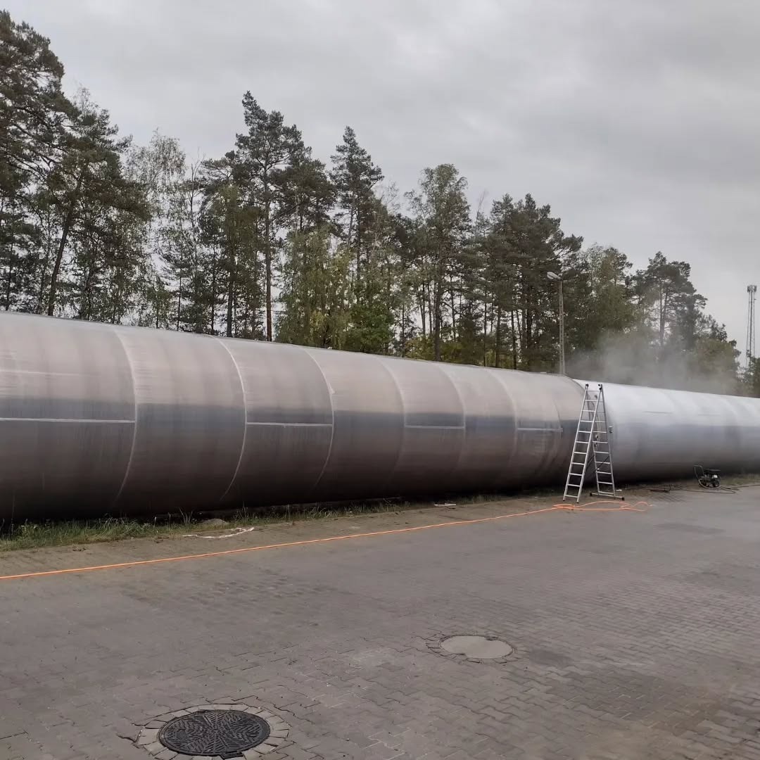 Długi, cylindryczny zbiornik z metalu częściowo umyty, widoczna drabina i sprzęt czyszczący, drzewa w tle. Część zbiornika jest jeszcze brudna.