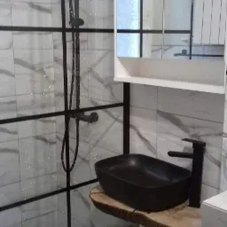 Nowoczesna łazienka z kabiną prysznicową typu walk-in z czarną armaturą, umywalką nablatową na blacie z plastra drewna i szafką z lustrem.