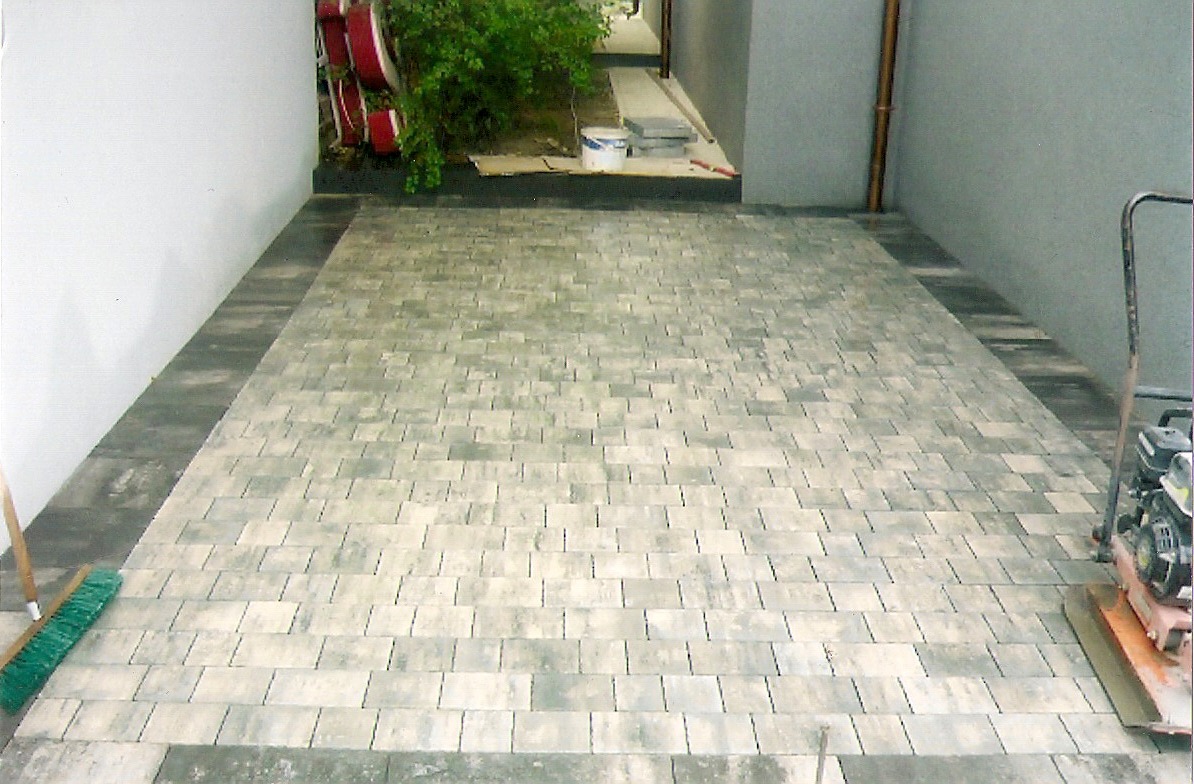 Ułożona kostka brukowa w odcieniach szarości i beżu, z widoczną ubijarką wibracyjną i szczotką do zamiatania, obramowana ciemniejszą kostką, z roślinnością w tle.