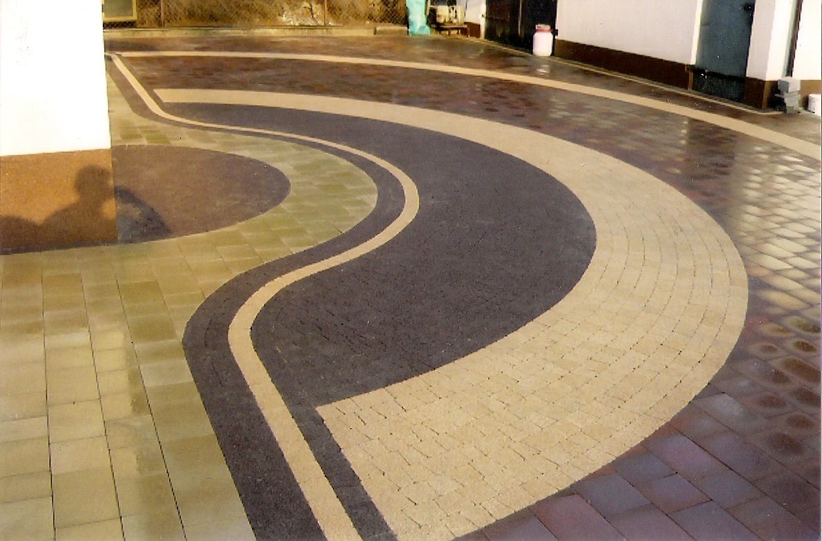 Wzorzysta nawierzchnia z kostki brukowej w odcieniach beżu, szarości i brązu, z zakrzywionymi liniami i okręgami, częściowo mokra, odbijająca światło.