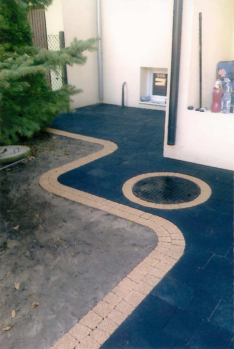 Ścieżka z ciemnej kostki brukowej obramowana jasną kostką w kształcie meandra i okręgu wokół studzienki, widok z góry.