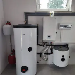 Instalacja kotła grzewczego i dwóch zbiorników ciepłej wody użytkowej w kotłowni, widok na białe urządzenia z czarnymi elementami na tle szarej ściany i podłogi, z oknem w tle.