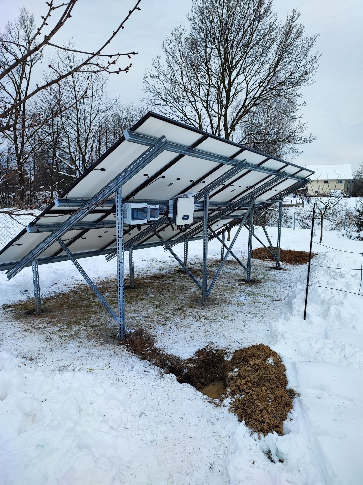 Montaż paneli fotowoltaicznych na metalowej konstrukcji w zimowym krajobrazie, widoczne wykopy w śniegu.