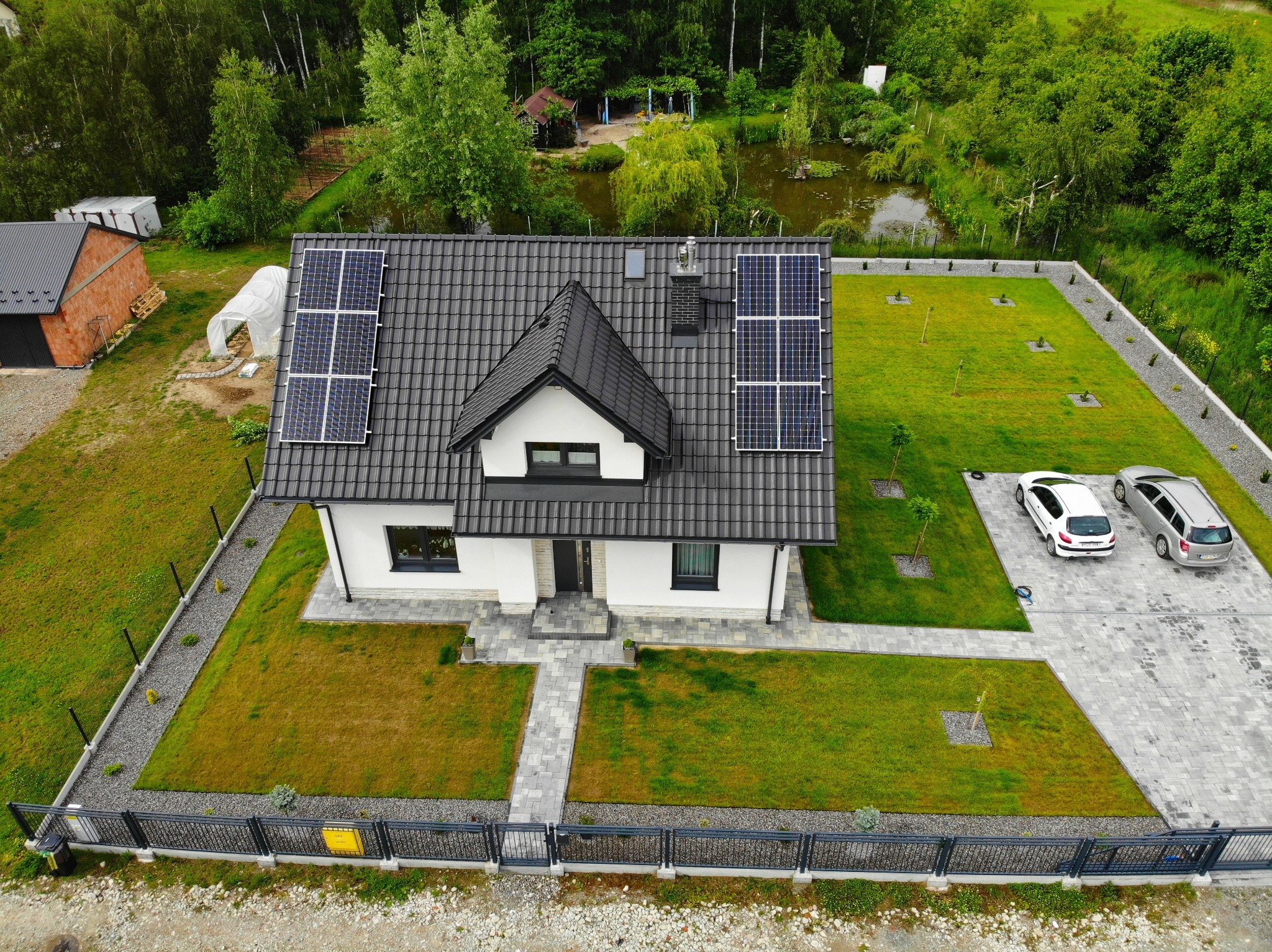 Dom jednorodzinny z panelami słonecznymi na dachu, widok z drona, zadbany trawnik i podjazd wyłożony kostką brukową, w tle staw i zieleń.