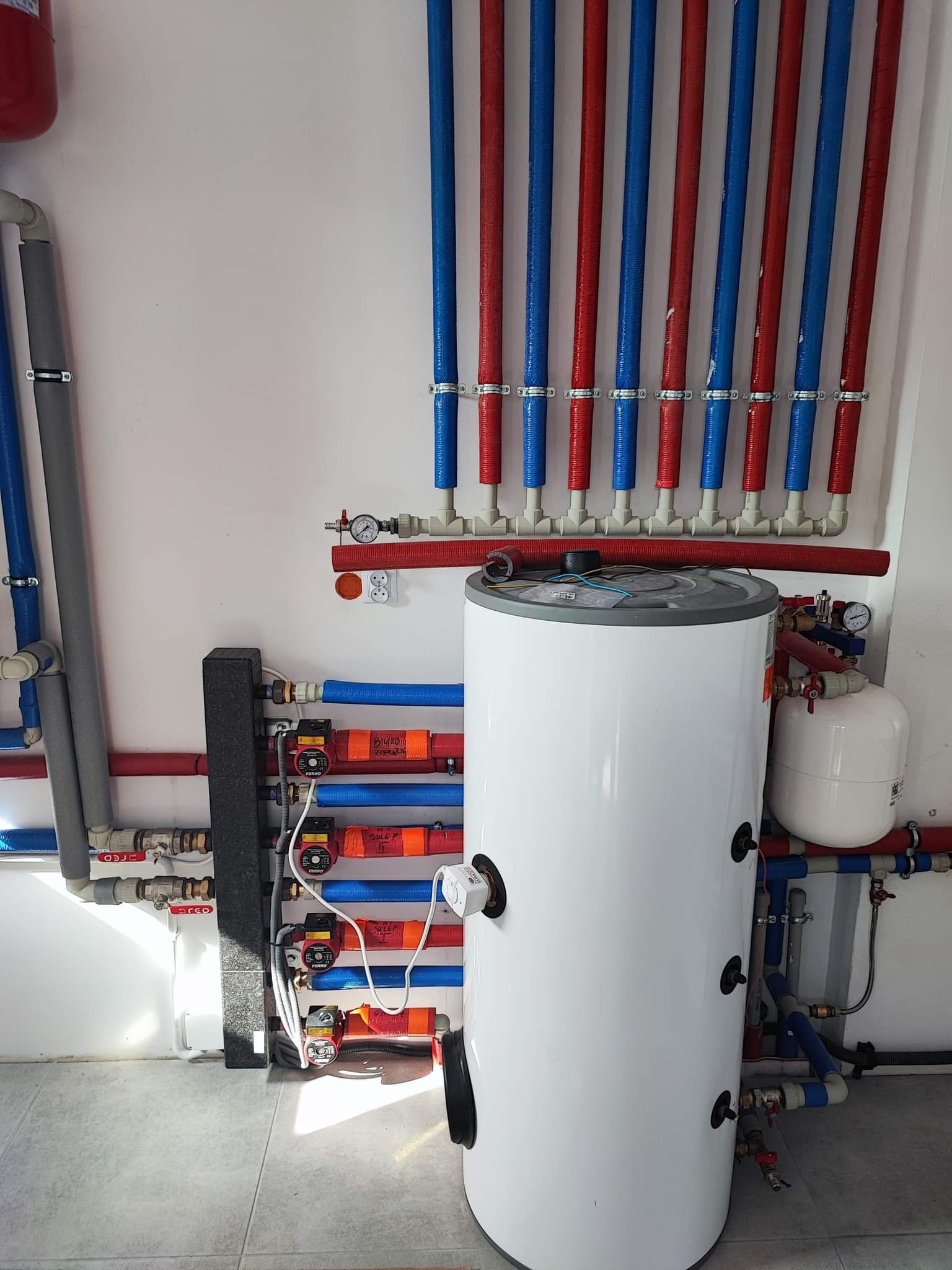 Instalacja grzewcza z białym zasobnikiem ciepłej wody, rurami w kolorach czerwonym i niebieskim, oraz pompami obiegowymi FERRO z pomarańczowymi etykietami, zamontowana na białej ścianie...
