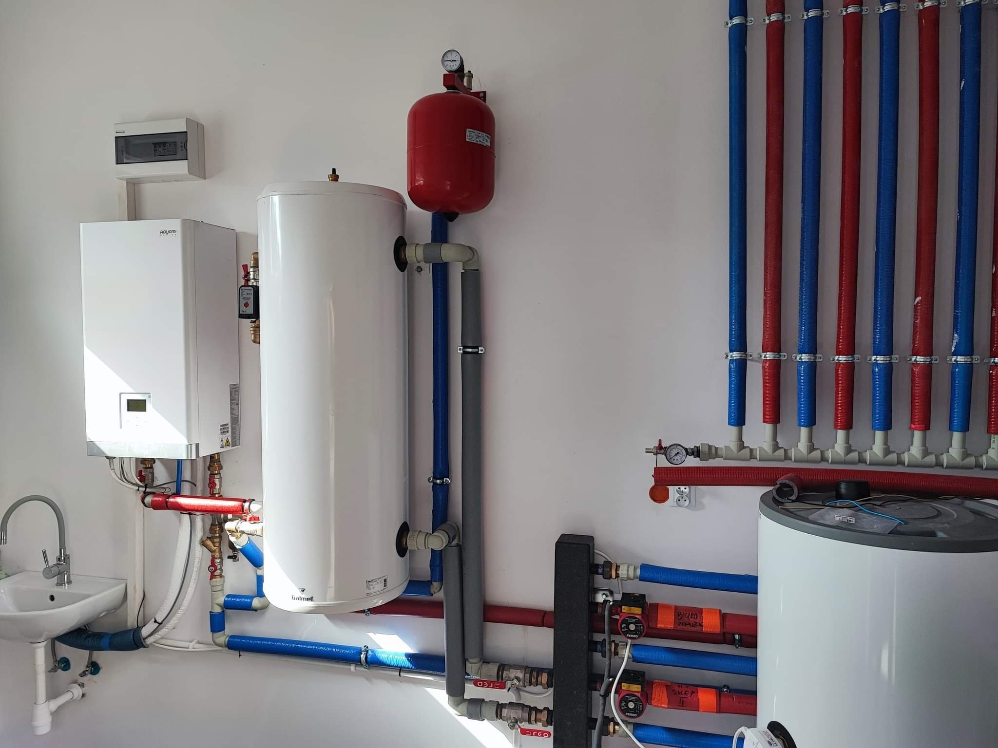 Instalacja centralnego ogrzewania z kotłem, zbiornikiem, rozdzielaczem i rurami w kolorach czerwonym i niebieskim, widoczna umywalka z kranem.