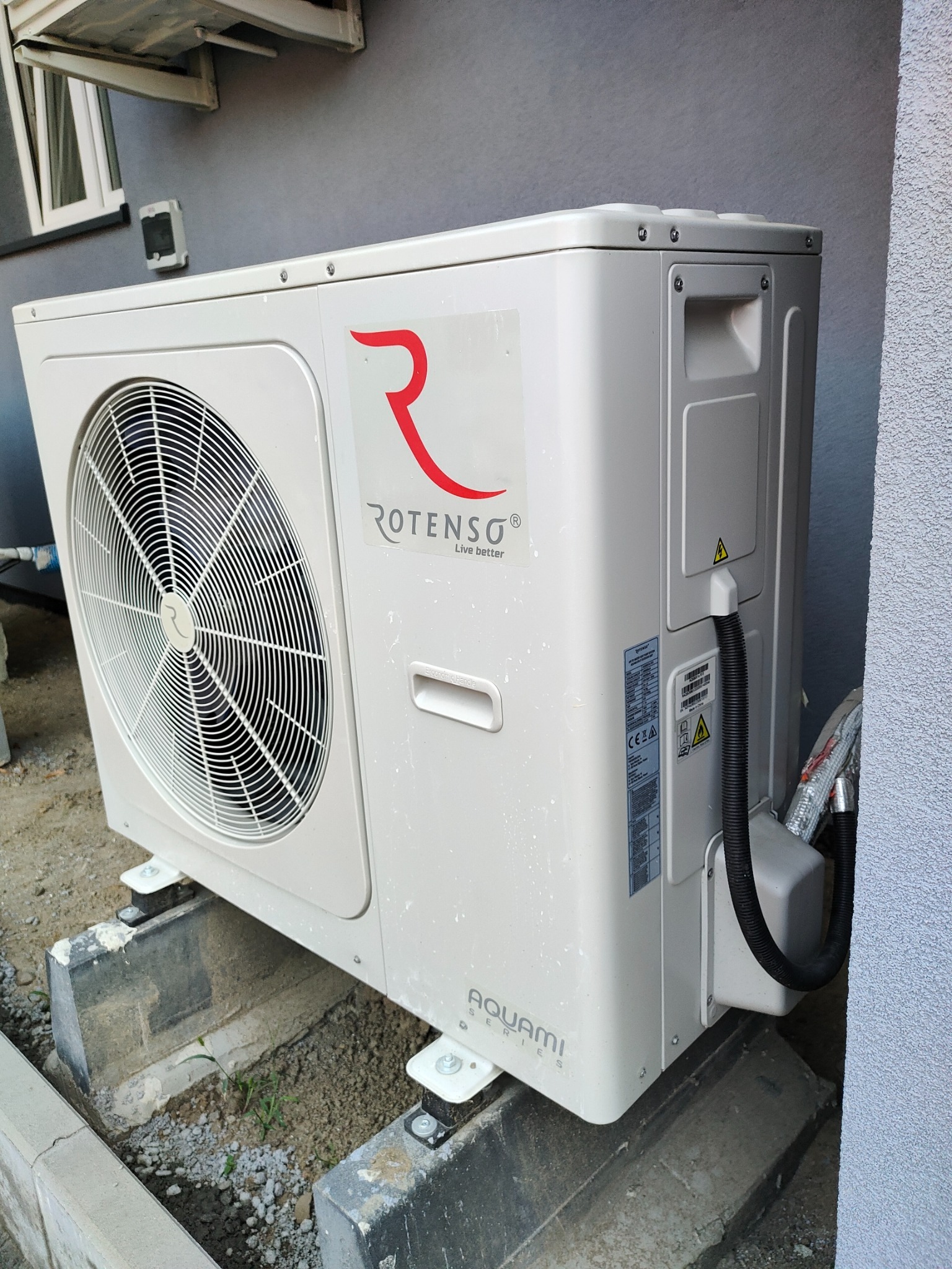 Zewnętrzna jednostka klimatyzacji Rotenso Aquami Series, zamontowana na betonowych podstawach przy szarej ścianie budynku.