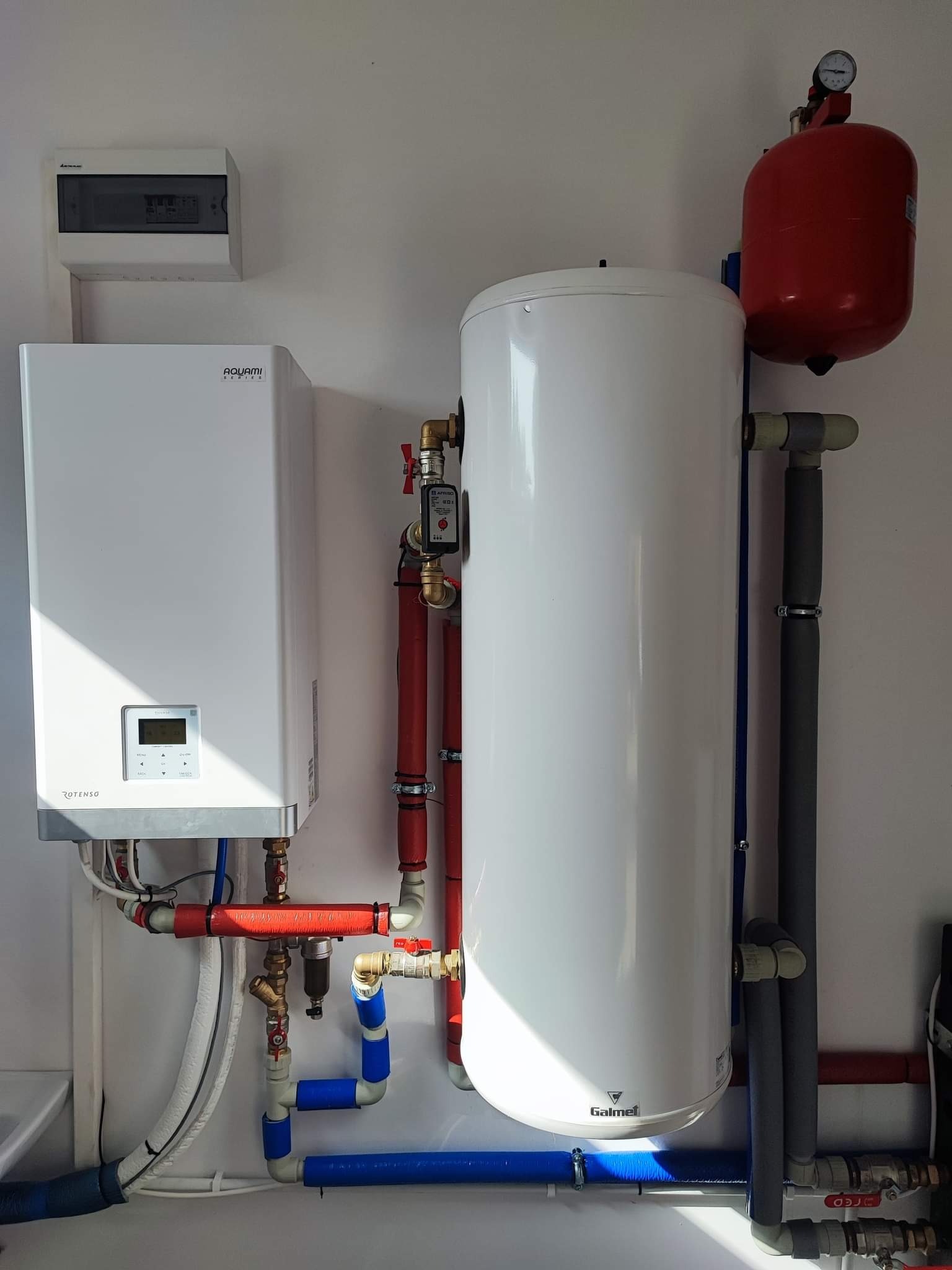 Biały bojler Galmet i piec Rotenso Aquami z podłączeniami rur miedzianych, plastikowych i izolowanych, zamontowane na białej ścianie, z widoczną skrzynką elektryczną u góry.