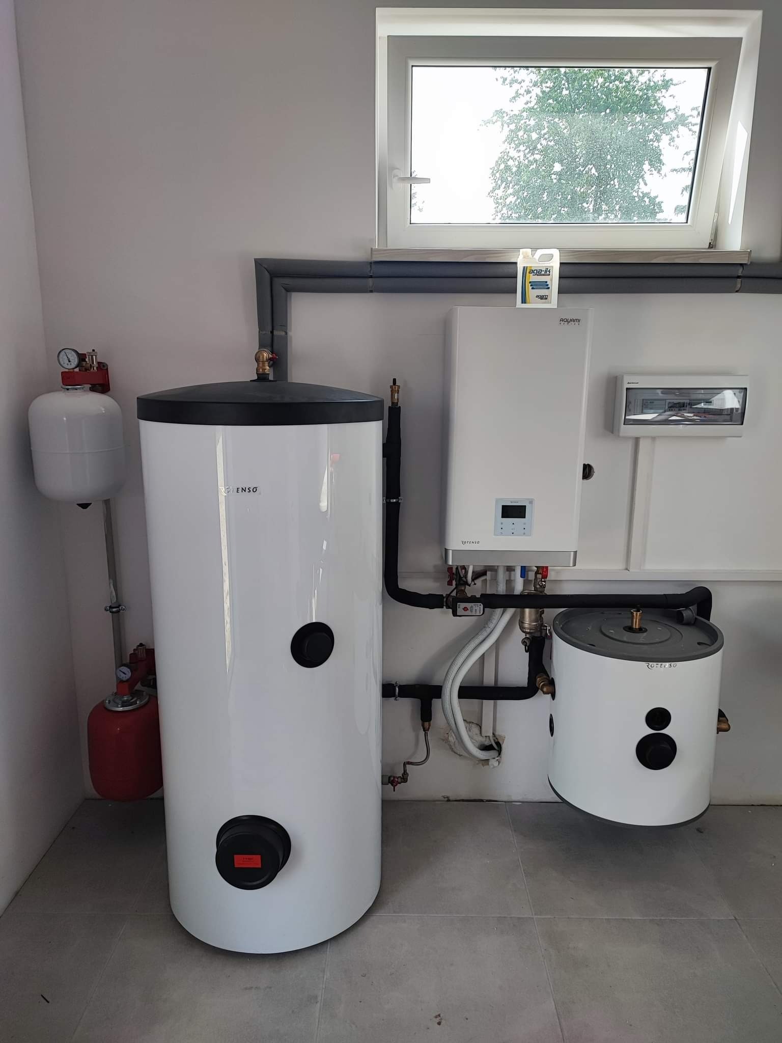 Instalacja kotła grzewczego i dwóch zbiorników ciepłej wody użytkowej w kotłowni, widok na białe urządzenia z czarnymi elementami na tle szarej ściany i podłogi, z oknem w tle.