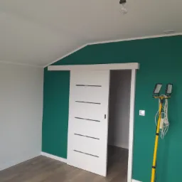 Pomalowany na zielono pokój z przesuwnymi białymi drzwiami z poziomymi wgłębieniami, jasnoszara ściana po lewej, drewnopodobna podłoga, żółty statyw z lampami doświetlającymi przy ścianie