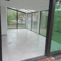 Nowoczesna, przeszklona weranda z białymi płytkami podłogowymi, widok z zewnątrz przez częściowo otwarte, przesuwne drzwi, widoczny fragment czerwonej kostki brukowej na zewnątrz.