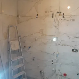 Montaż płytek imitujących marmur na ścianie łazienki, widoczne krzyżyki dystansowe, poziomica laserowa i drabina aluminiowa.