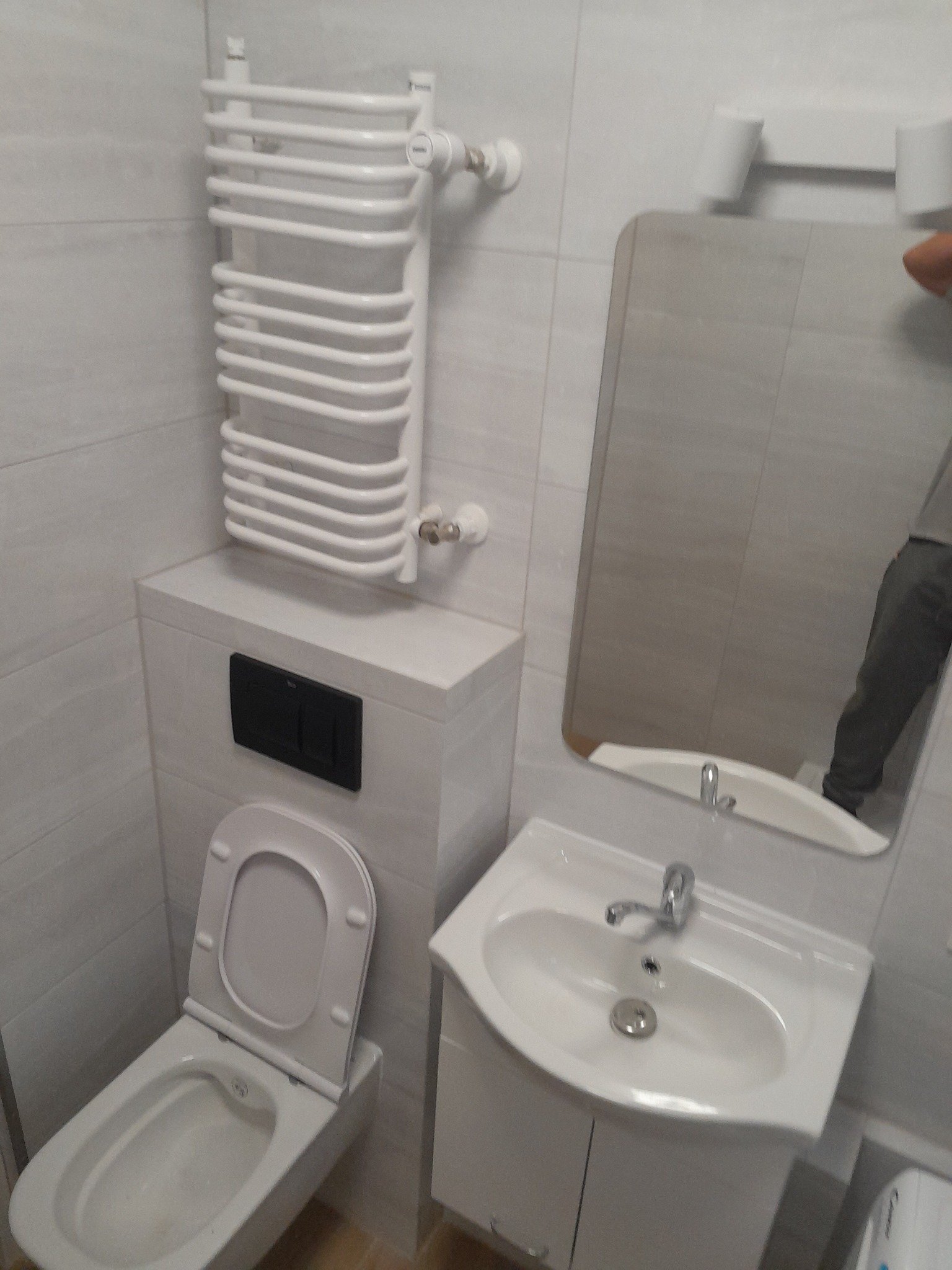 Biała łazienka z wiszącą toaletą z czarnym przyciskiem spłukiwania, grzejnikiem drabinkowym i umywalką z lustrem, odbicie części osoby.