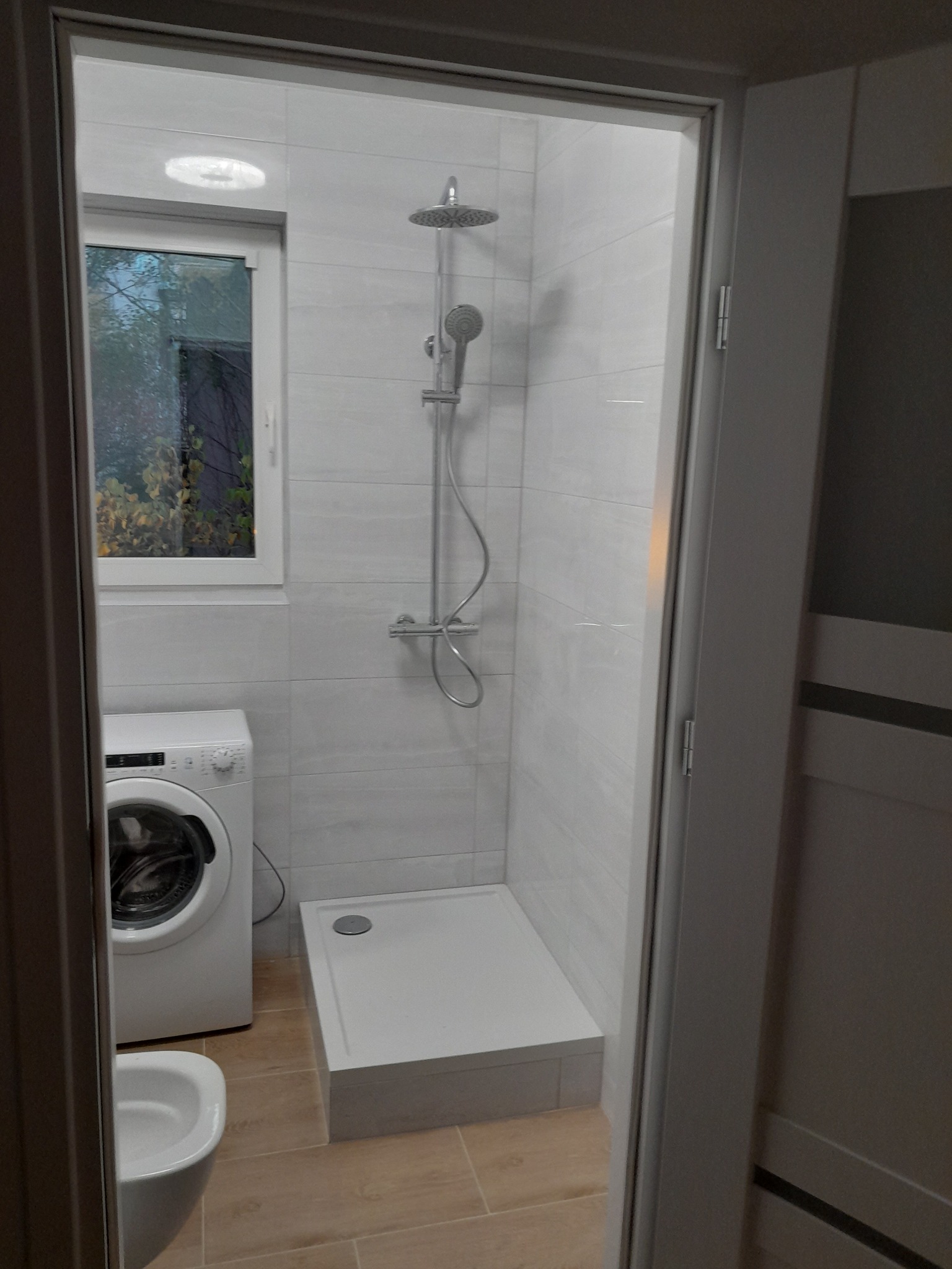 Nowoczesna kabina prysznicowa z deszczownicą, jasne płytki na ścianach, beżowa podłoga imitująca drewno, pralka i toaleta w tle, widok zza framugi drzwi.