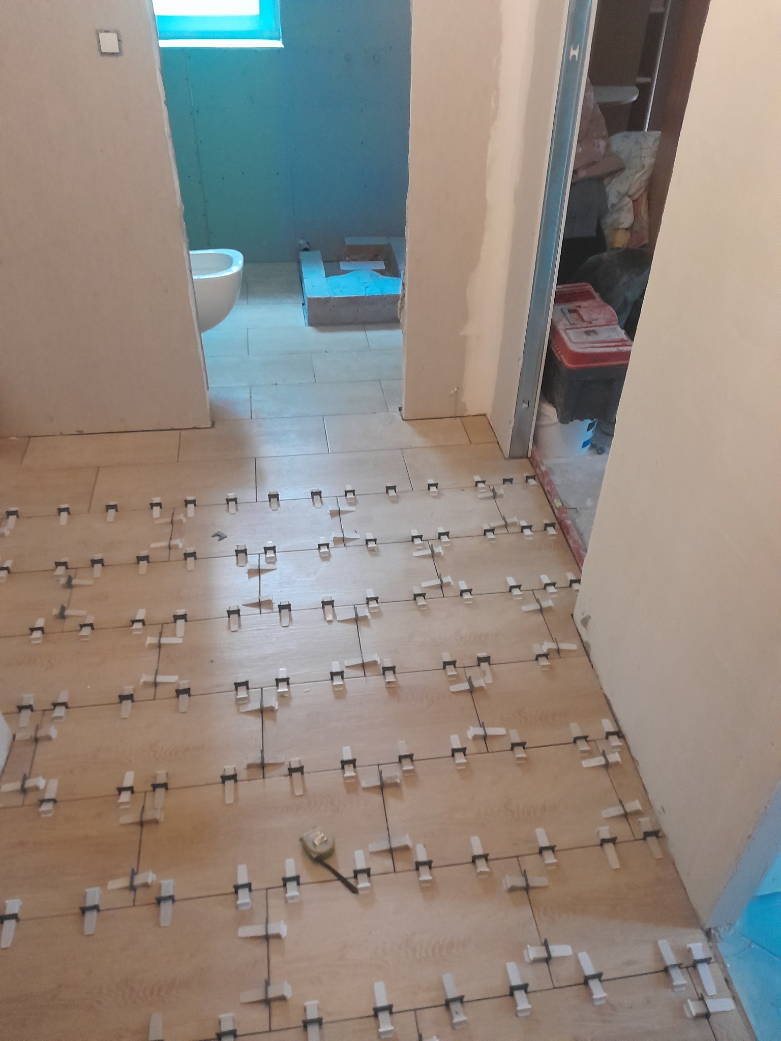 Układanie podłogi z płytek imitujących drewno w łazience, z użyciem klipsów poziomujących, widoczny fragment toalety i narzędzia pomiarowe.