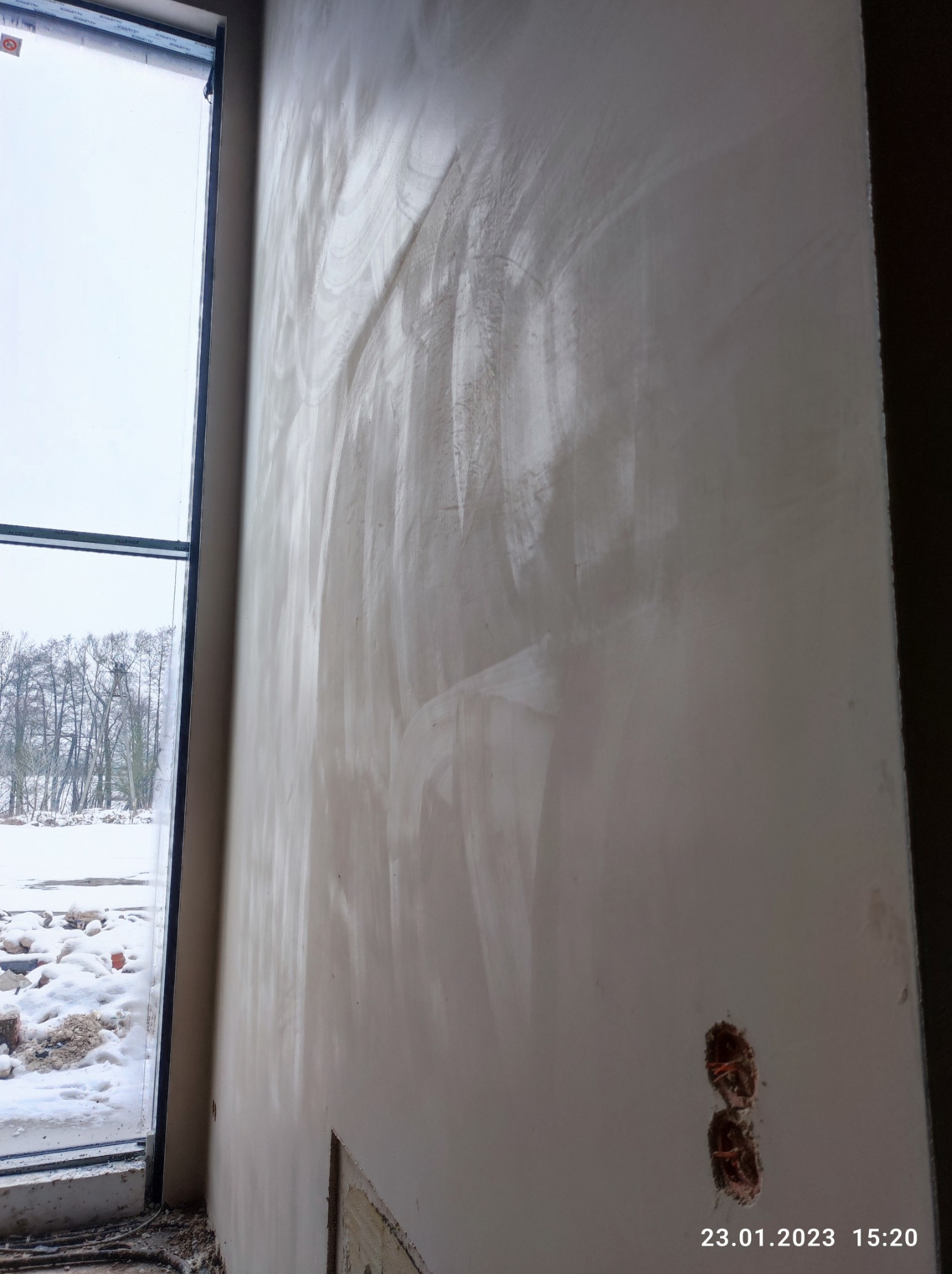 Świeżo nałożony tynk maszynowy na ścianie wewnętrznej, widoczne ślady po maszynie, niezakończone prace wykończeniowe, gniazdka elektryczne bez osprzętu, okno z widokiem na zimowy krajobraz.