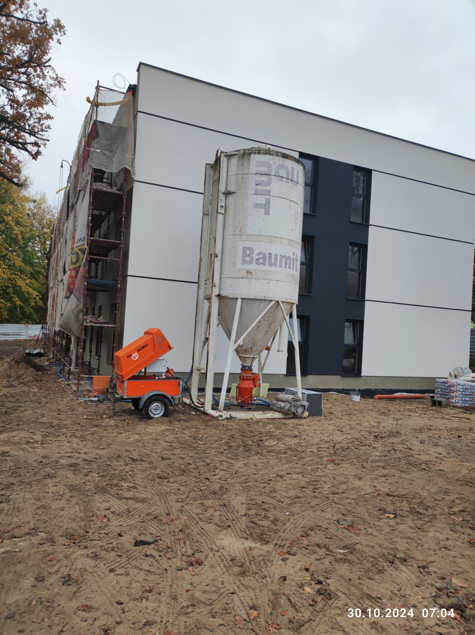 Silos z zaprawą Baulit i agregat tynkarski Silomat PFT na placu budowy, widoczne rusztowania przy ocieplonym budynku.