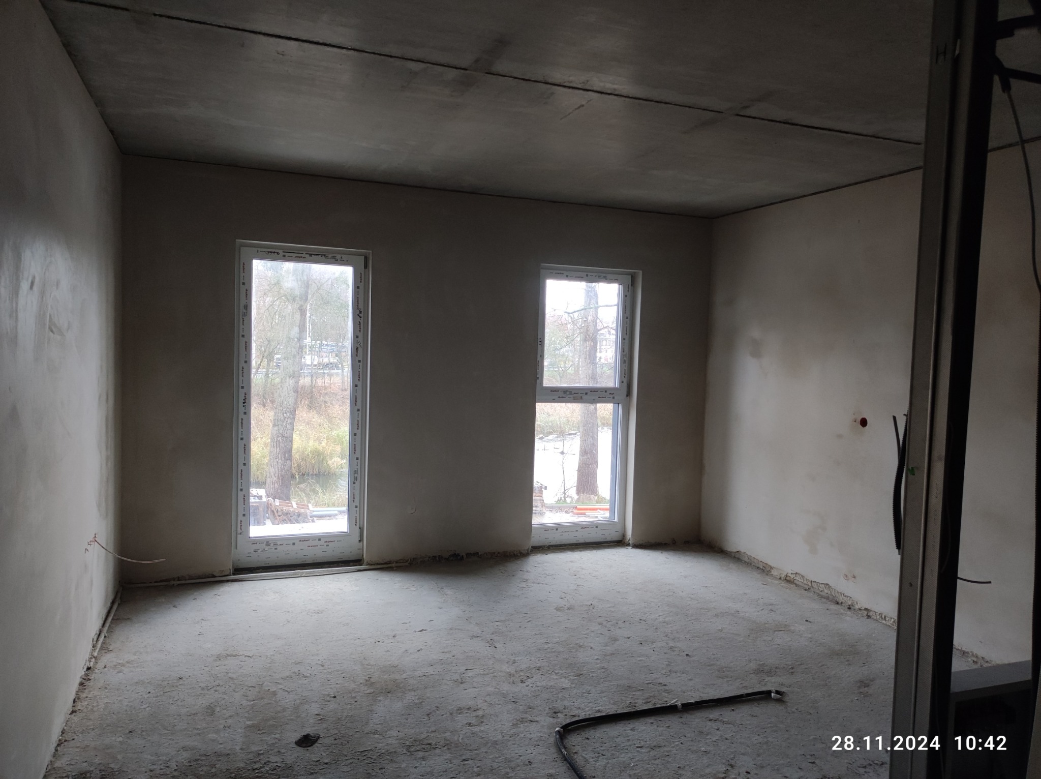 Surowe wnętrze po tynkowaniu maszynowym, widoczne dwa okna z widokiem na zewnątrz, betonowa podłoga i ściany z szarym tynkiem, instalacja elektryczna wystająca ze ściany, fragment metalowej...