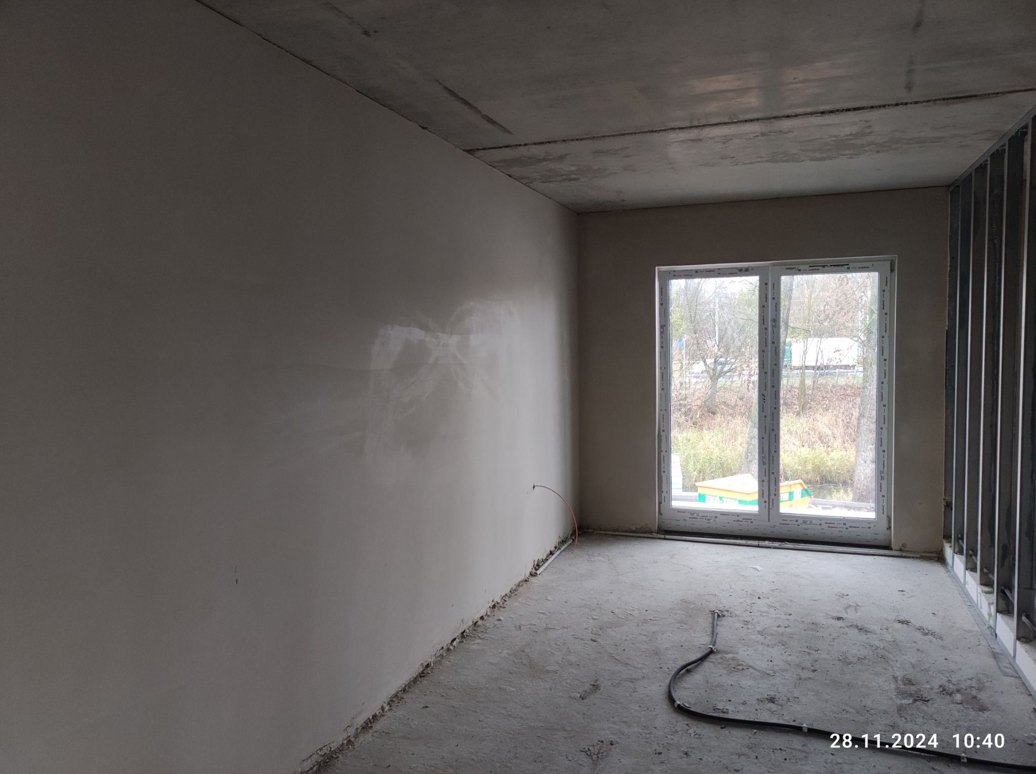 Wnętrze surowego pomieszczenia po nałożeniu gładzi gipsowej na ściany, z widoczną instalacją elektryczną wystającą ze ściany i profilem konstrukcyjnym przy ścianie z oknem.