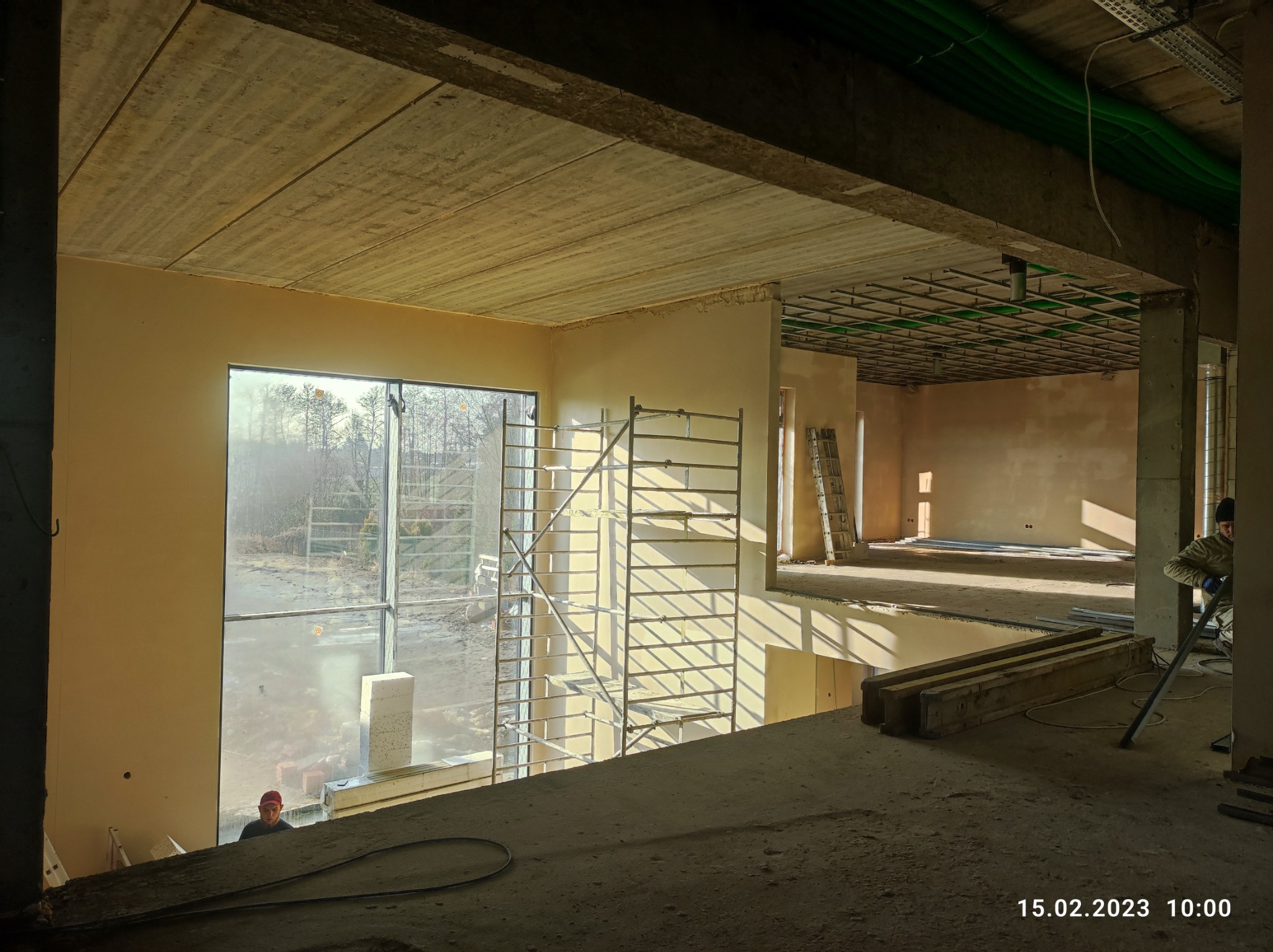 Wnętrze budynku w trakcie prac wykończeniowych; widoczne ściany po nałożeniu gładzi, rusztowania przy oknie, instalacje pod sufitem i pracownik w czerwonej czapce w tle.