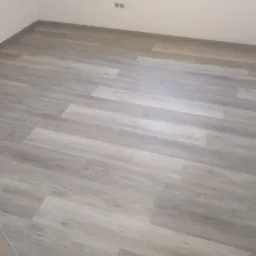 Ułożone panele podłogowe w odcieniach szarości w narożnym pokoju, widoczne gniazdka elektryczne na ścianie.