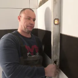 Mężczyzna w ubraniu roboczym sprawdza poziomicy ułożenie białych i ciemnych płytek na ścianie w trakcie prac wykończeniowych.