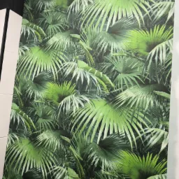 Ściana pokryta tapetą z motywem gęstych, zielonych liści palmowych, tworzących iluzję tropikalnego lasu, z widocznymi fragmentami białej listwy przypodłogowej i czarnego sufitu.