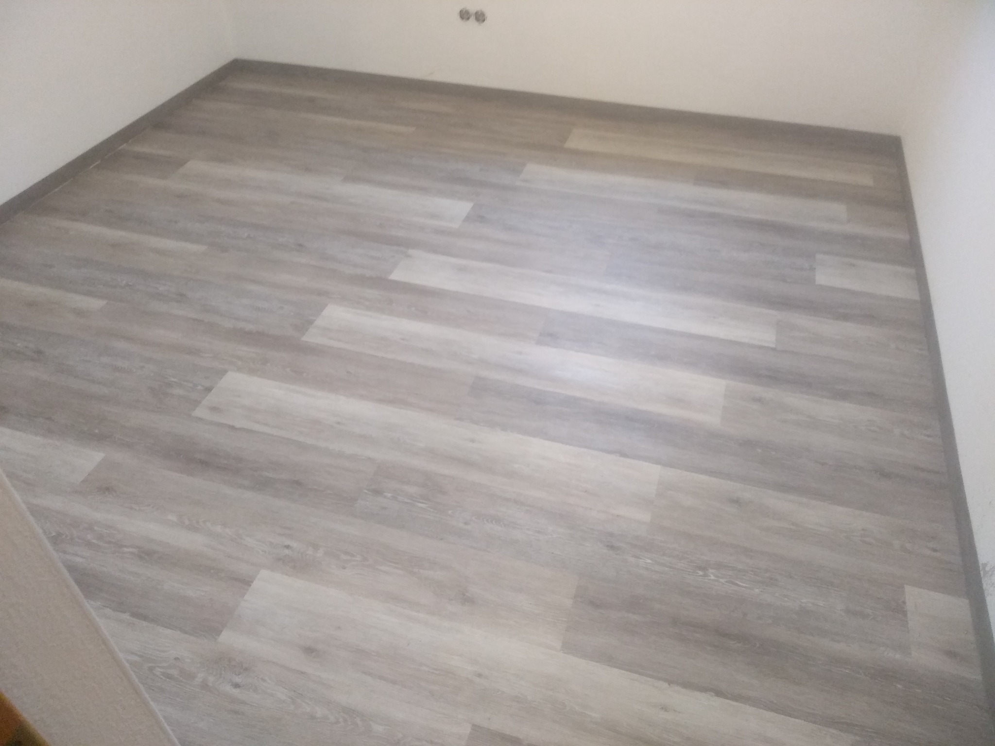 Nowo ułożone panele podłogowe w odcieniach szarości, widoczne gniazdka elektryczne na ścianie, świeżo wykończone pomieszczenie.