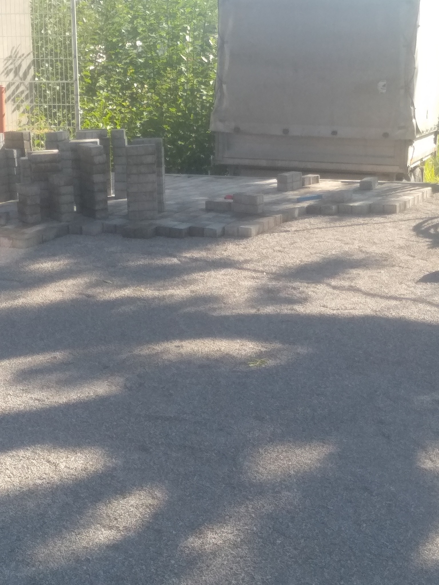 Układanie szarej kostki brukowej na podsypce, widoczne stosy kostki i częściowo ułożony fragment nawierzchni, w tle zieleń i fragment ciężarówki.