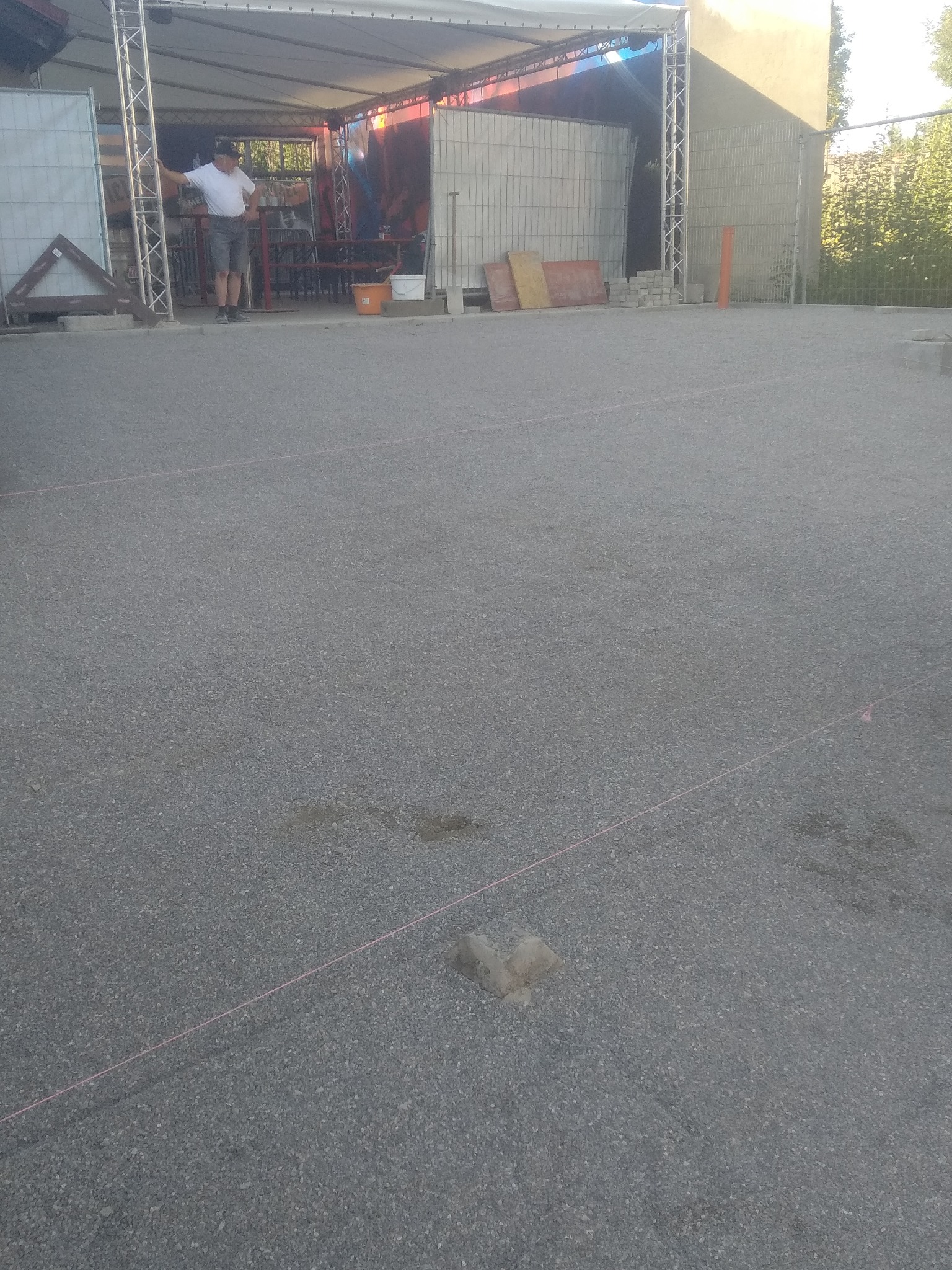 Przygotowanie terenu pod budowę: widoczny fragment utwardzonej nawierzchni z wyznaczonymi liniami, w tle mężczyzna oparty o konstrukcję z metalowych rusztowań, elementy budowlane i wiadra.