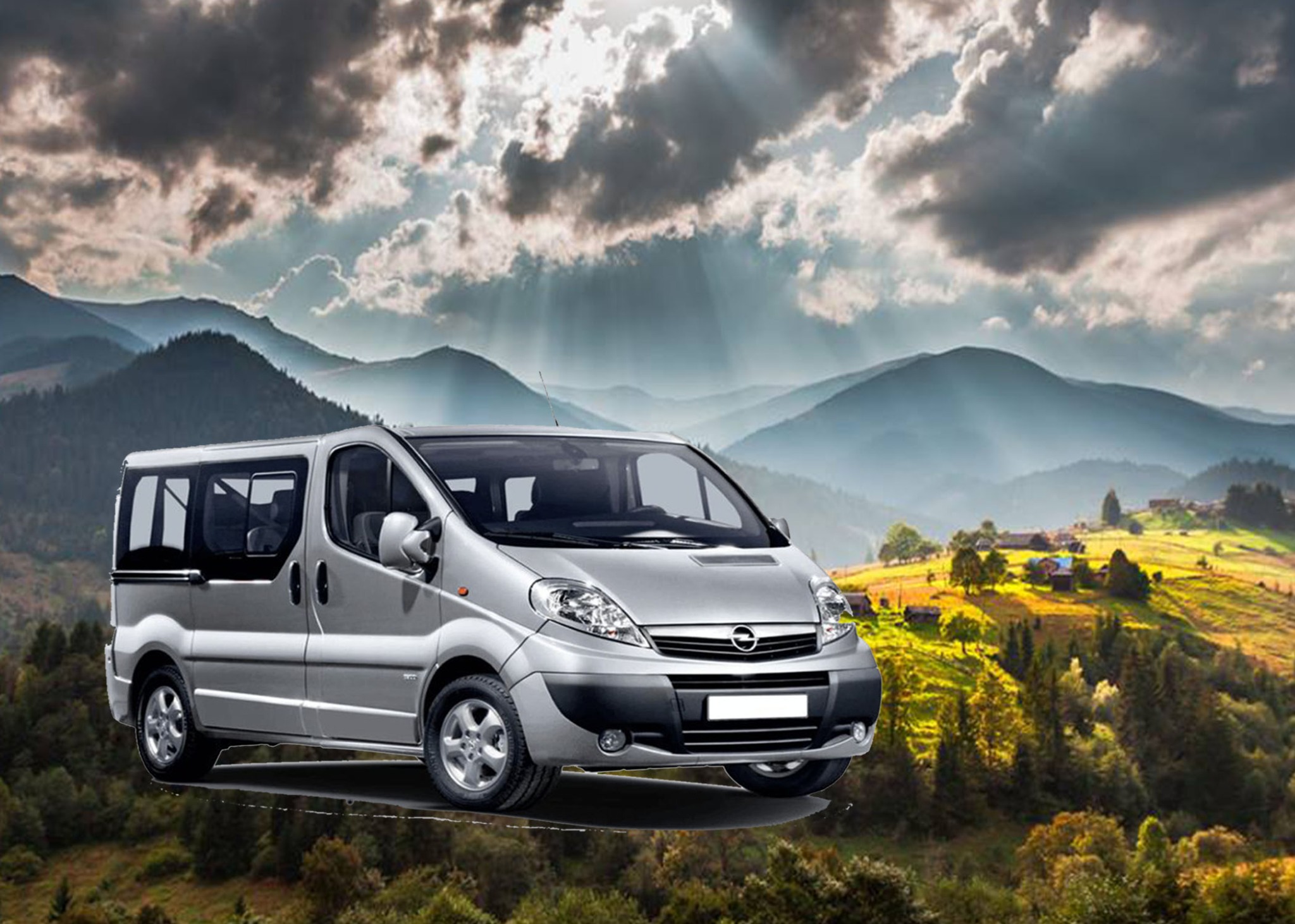 Srebrny van na tle górskiego krajobrazu z promieniami słońca przebijającymi się przez chmury.