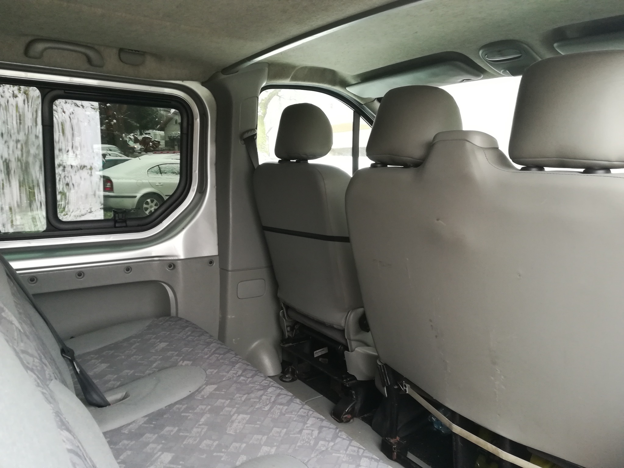 Wnętrze busa z szarymi fotelami i wzorzystą tapicerką, widok z tyłu na przednie siedzenia i okno z widokiem na zaśnieżony samochód.