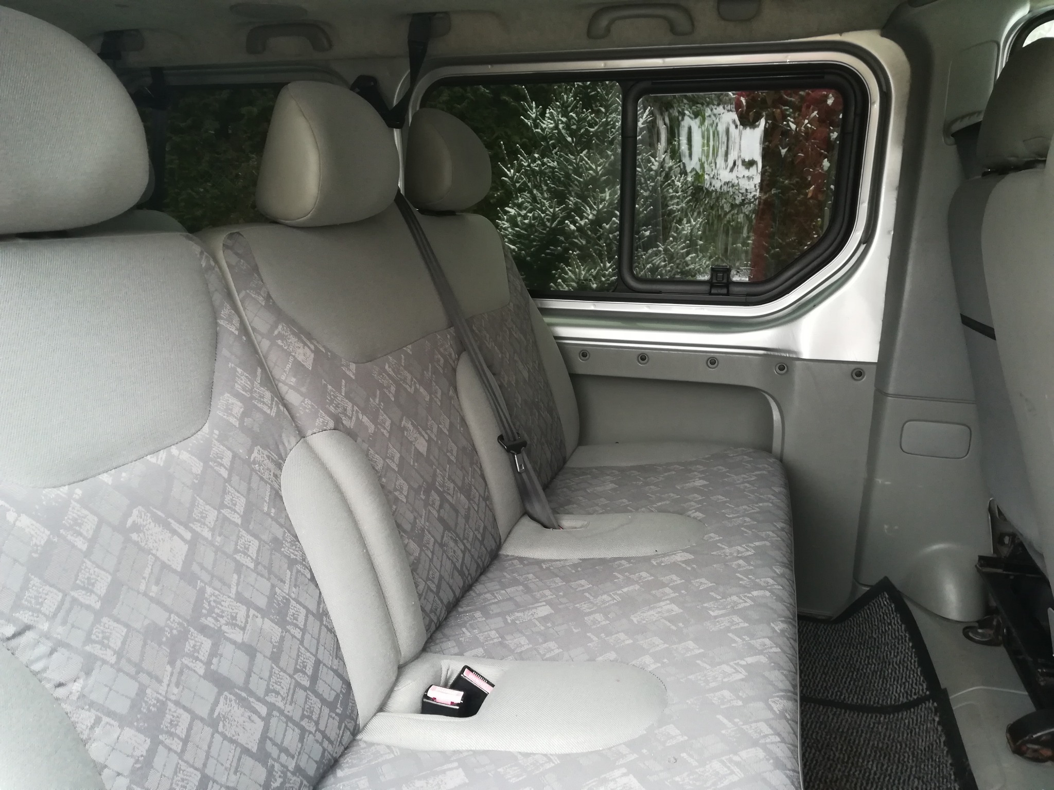 Wnętrze srebrnego busa: tylne siedzenia z szarą tapicerką w geometryczne wzory, widok przez okno na zieleń.