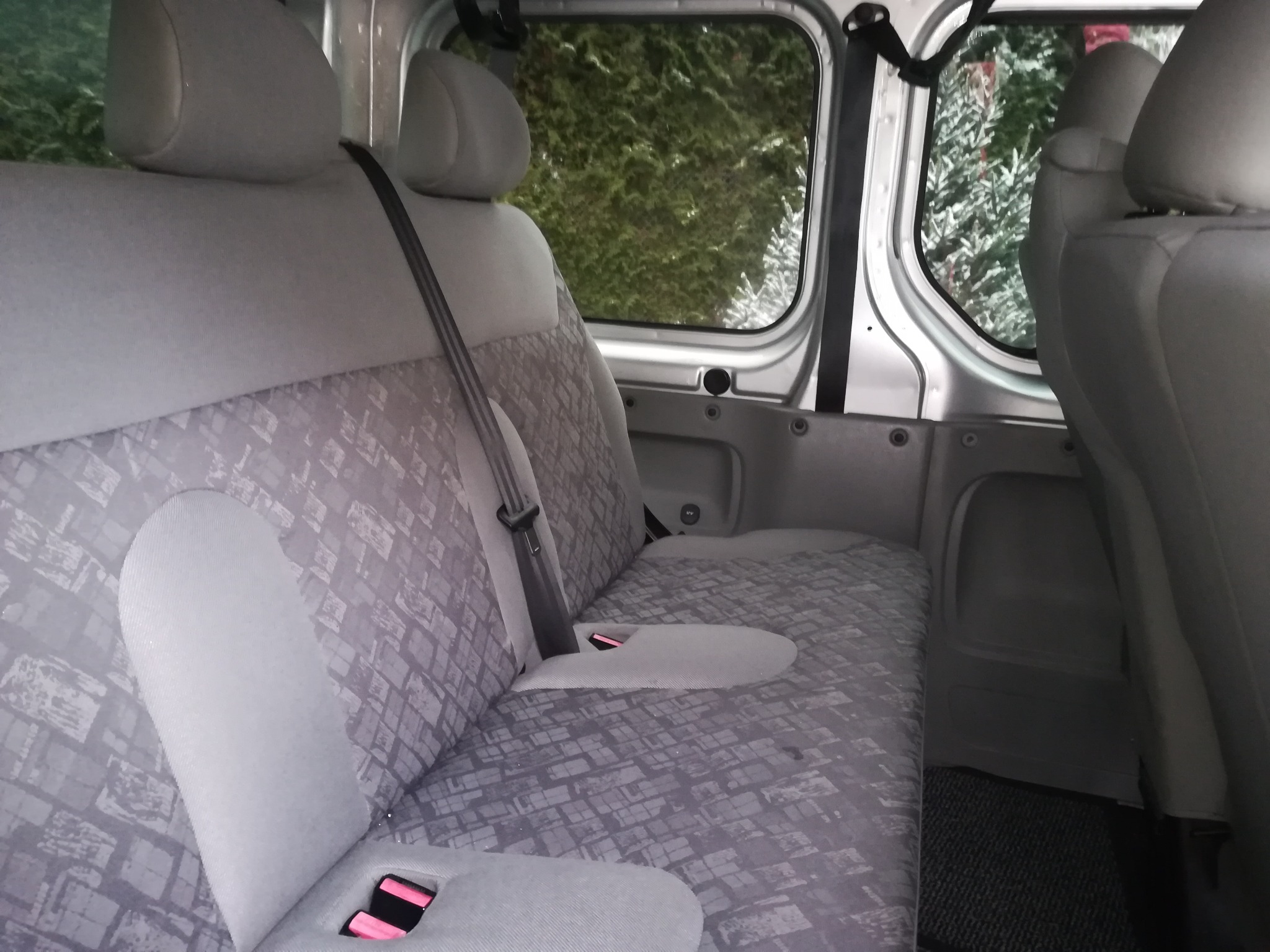Wnętrze srebrnego busa z szarą tapicerką w geometryczny wzór, widoczne fotele pasażerów i zieleń za oknem.