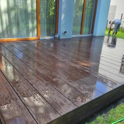 Zloty Ogród - Mokry taras kompozytowy przy domu z dużymi oknami, osoba czyści powierzchnię. Widoczne krople wody i odbicia. Zastosowano deski w odcieniach brązu.