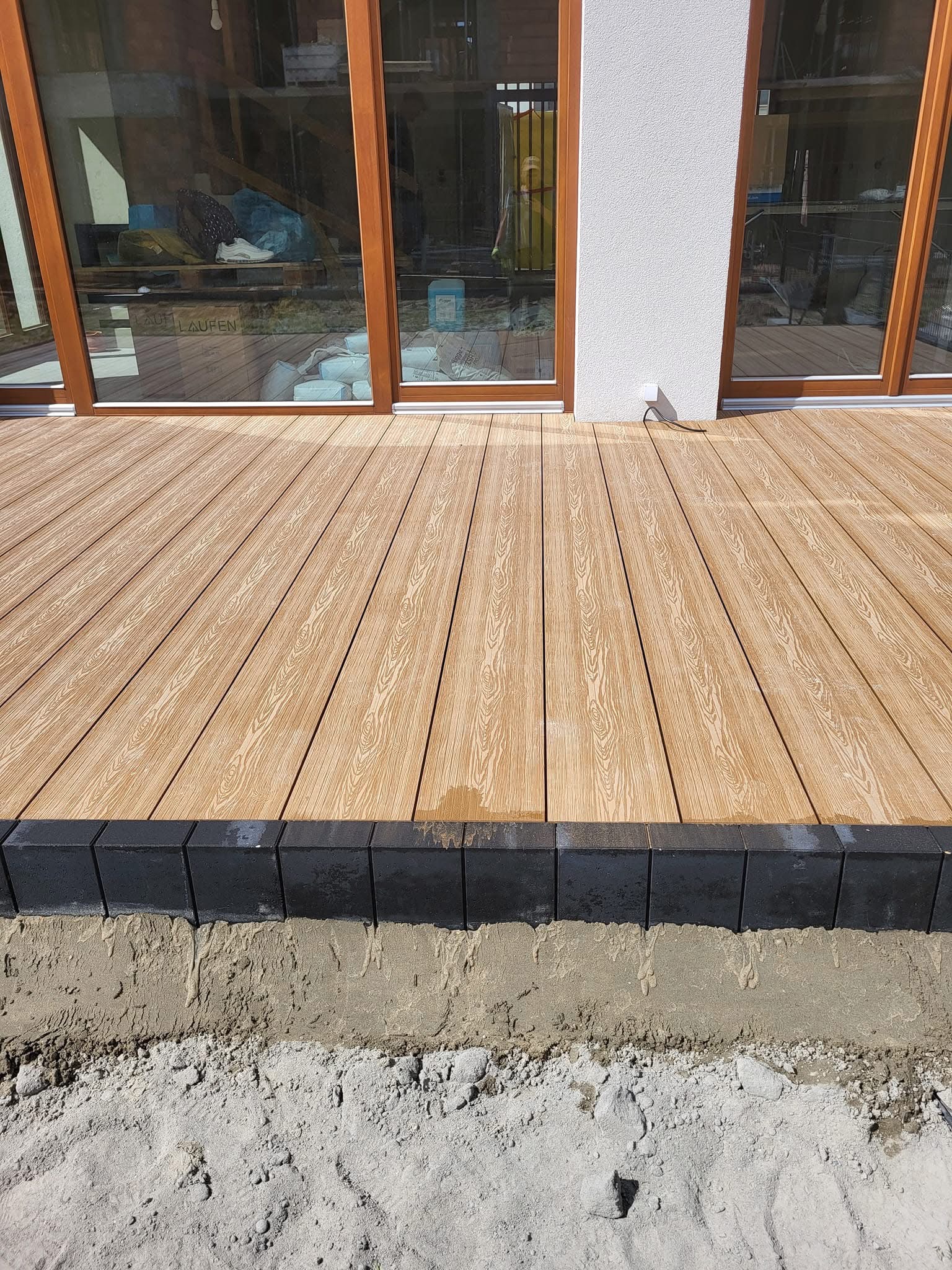 Jasny taras kompozytowy z czarną obwódką z kostki, widok z bliska na etapie budowy, z piaskiem i ziemią w tle. Duże okna tarasowe w tle.