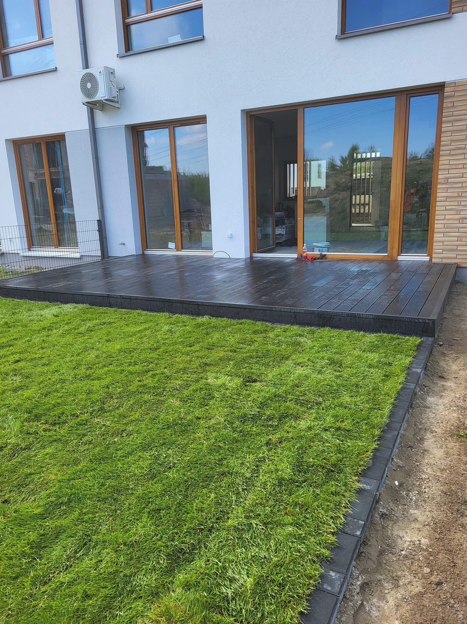 Nowoczesny taras z ciemnego drewna przylegający do domu, otoczony świeżo ułożoną, soczystą trawą. Widoczne detale wykończenia i granica trawnika z kostki brukowej.