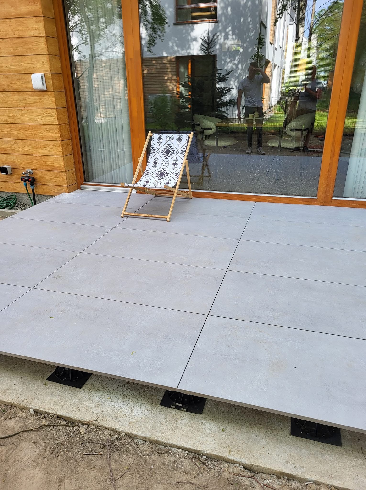 Jasnoszary taras z płytek na wspornikach, z drewnianym krzesłem plażowym. Widoczne duże okno tarasowe i fragment drewnianej elewacji budynku.