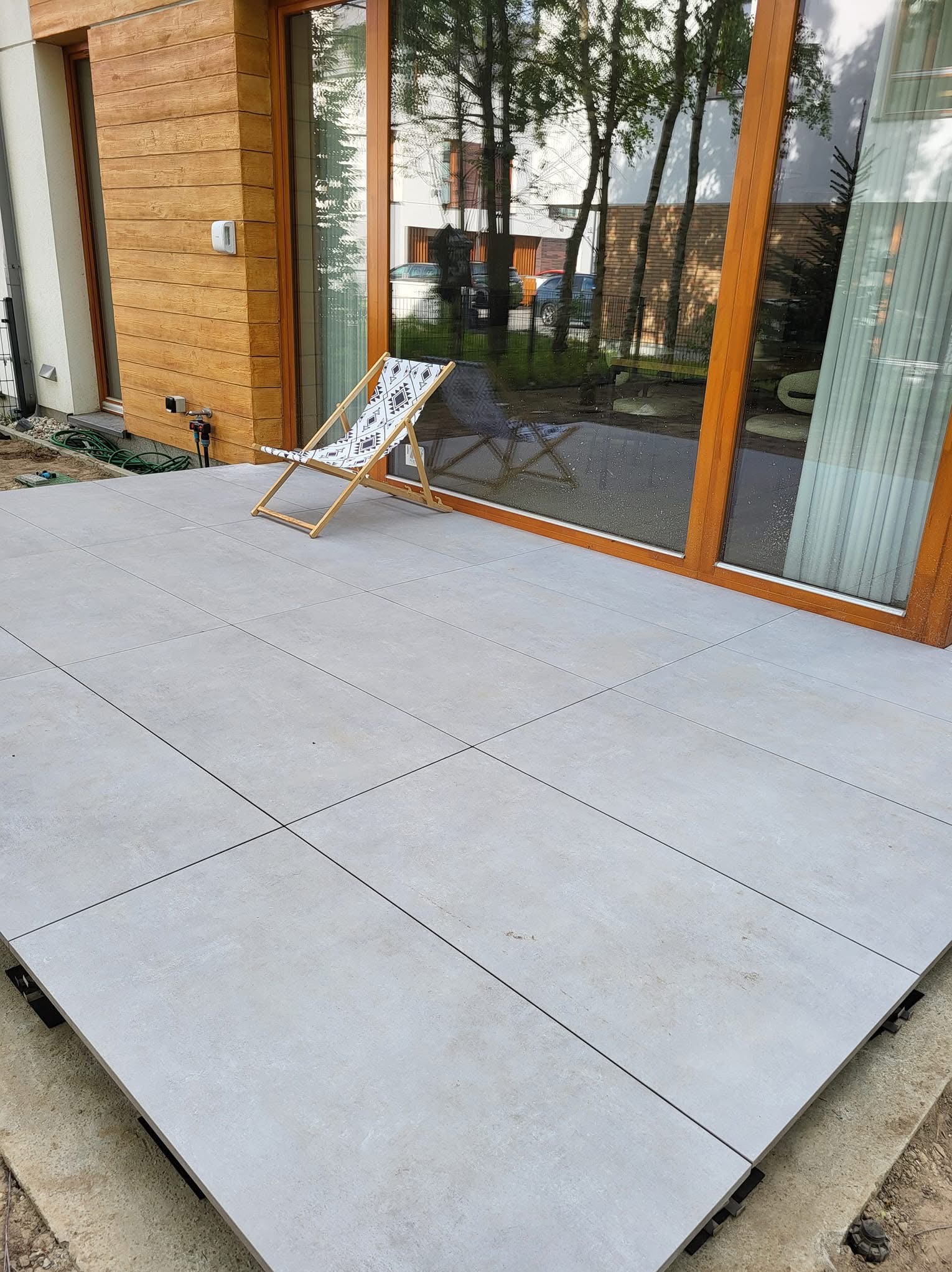 Jasnoszary taras z płytek ceramicznych przy domu z drewnianą elewacją. Na tarasie leżak z geometrycznym wzorem. Duże okno tarasowe z drewnianą ramą.