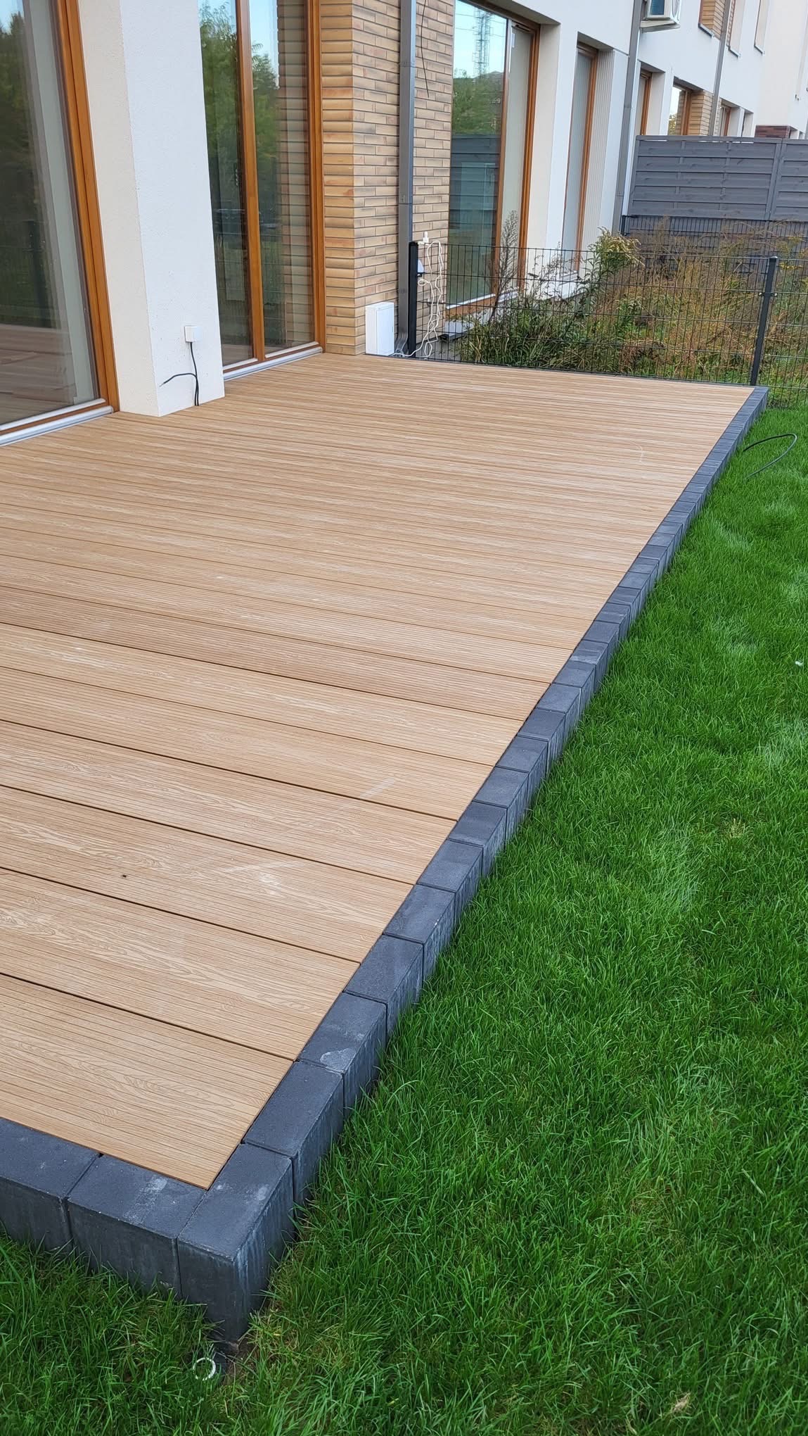Jasny taras kompozytowy z szarą obwódką z kostki brukowej, przylegający do budynku i trawnika. Widok z góry, oświetlenie dzienne.