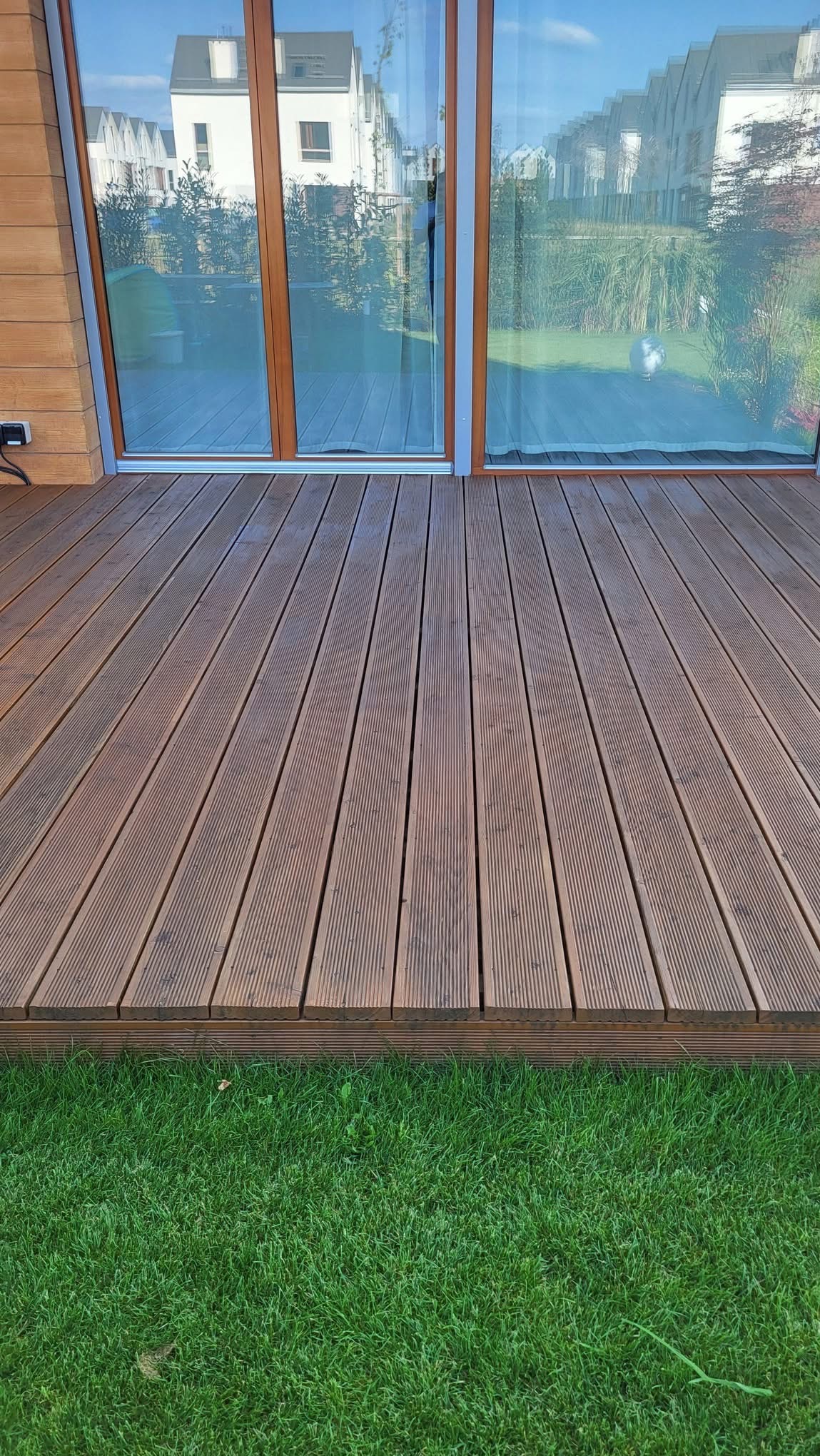 Nowoczesny taras z desek kompozytowych w odcieniach brązu, przylegający do budynku z dużymi oknami, otoczony zielonym trawnikiem. Widok z niskiej perspektywy.