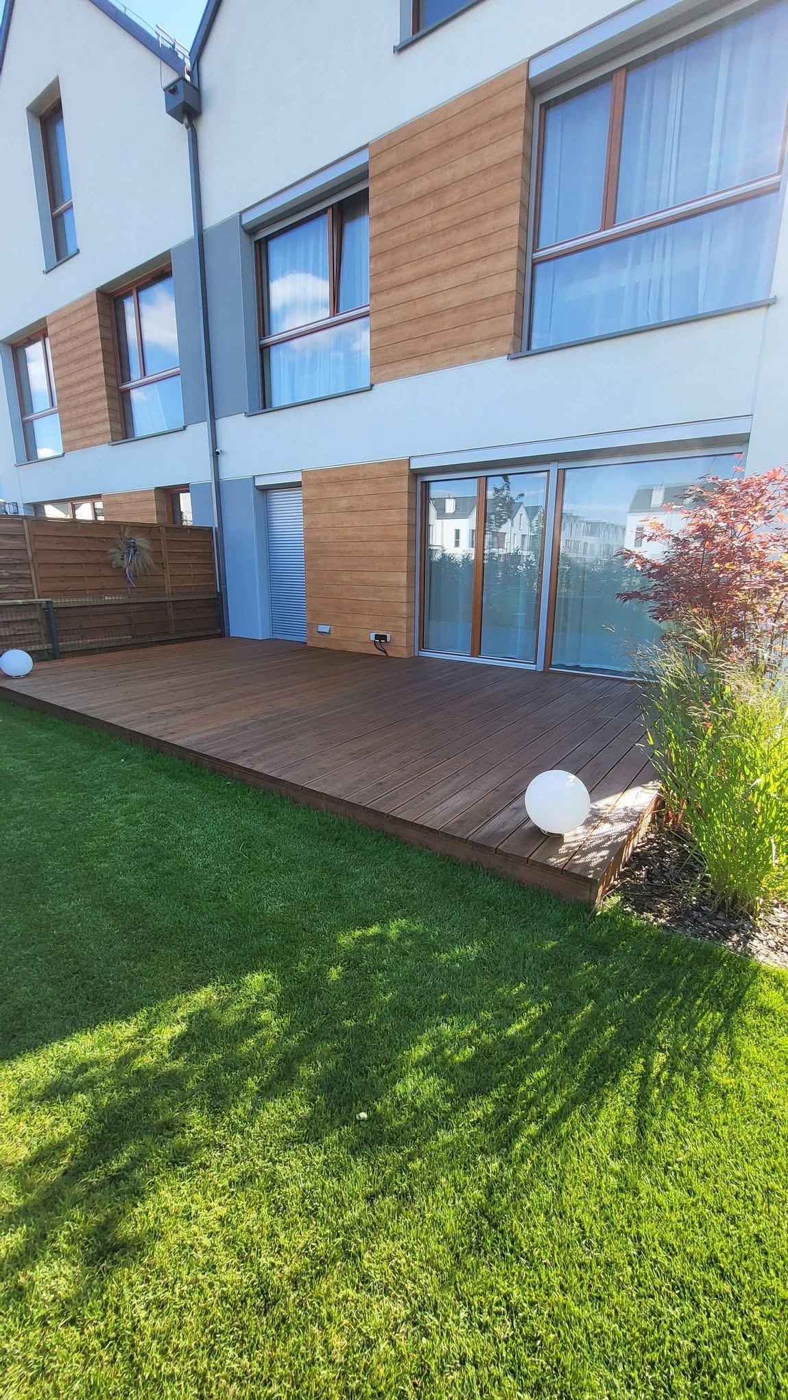 Nowoczesny taras drewniany z oświetleniem kulistym, przylegający do budynku z drewnianą elewacją i zadbanym trawnikiem. Widoczne rośliny ozdobne.