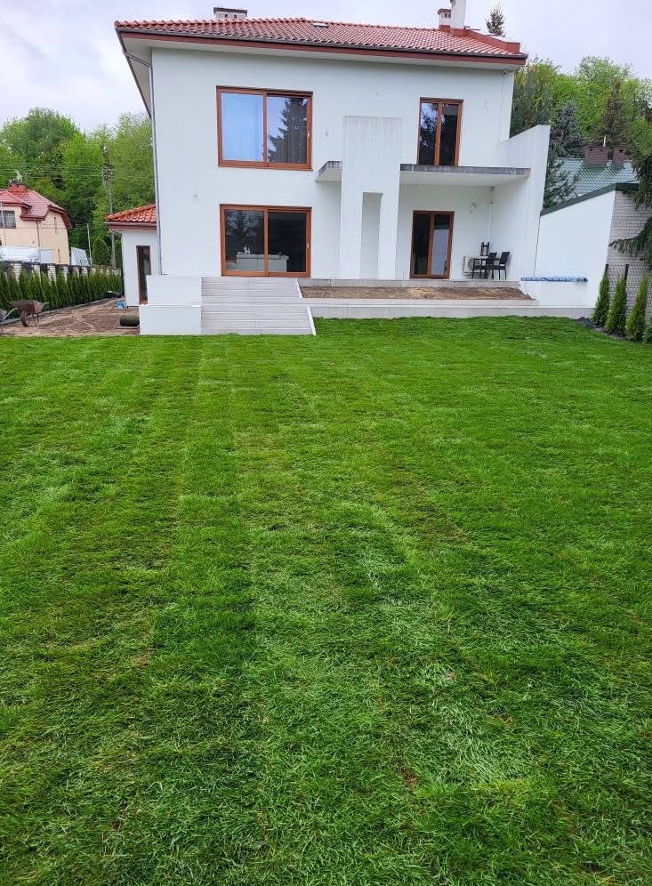 Nowoczesny dom z zadbanym trawnikiem, świeżo po koszeniu. Widoczne pasy po kosiarce, schludne otoczenie domu i elementy architektury ogrodowej.