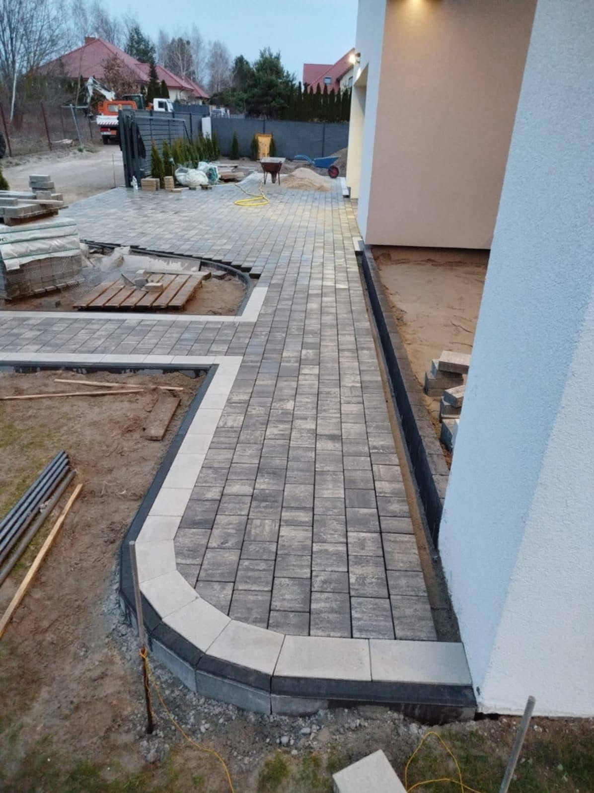 Ścieżka z kostki brukowej w odcieniach szarości, obrzeża z jasnych kostek. Częściowo ułożona, widoczne materiały i narzędzia. Tło: dom jednorodzinny, elementy ogrodu.